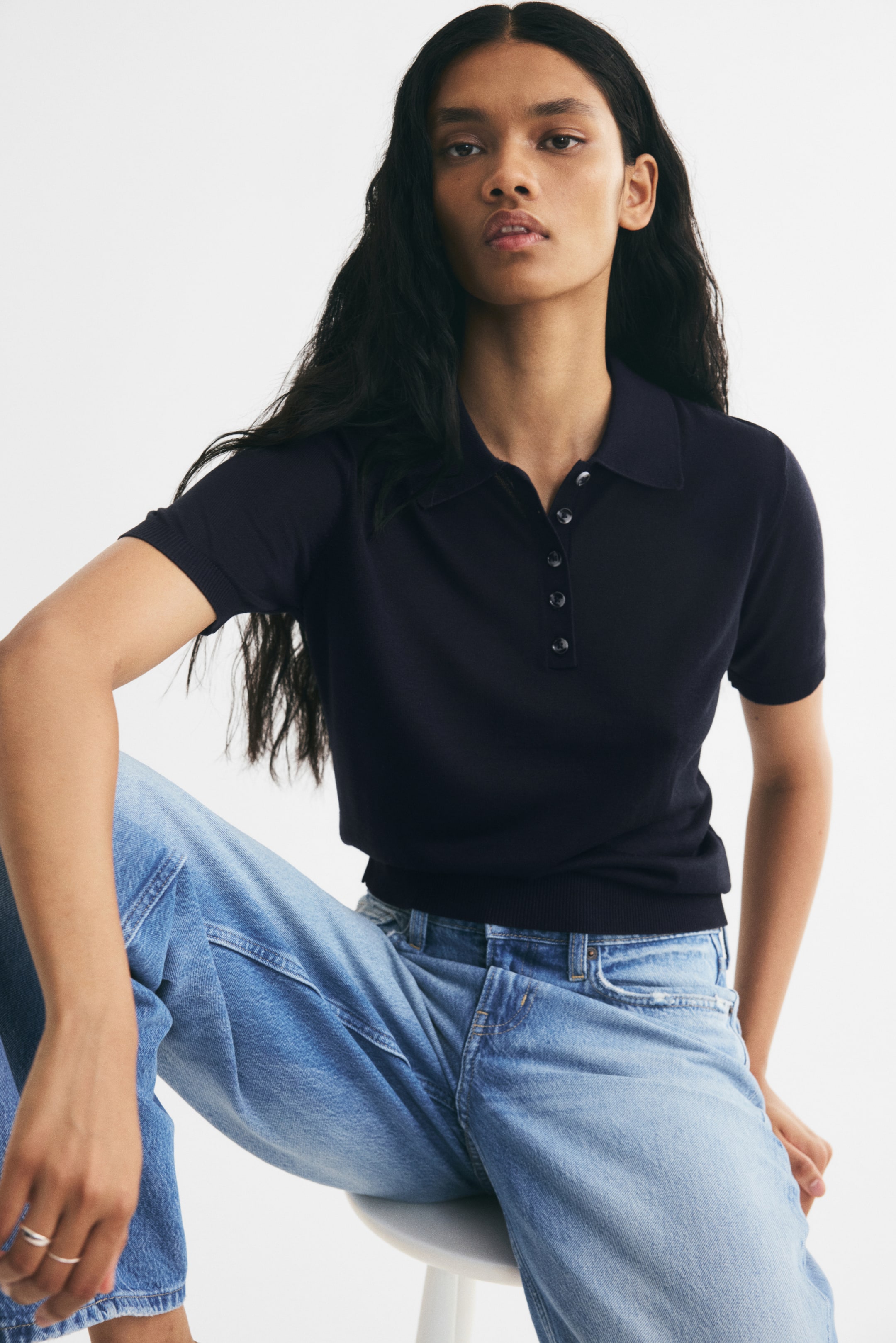 View larger image: A dark navy blue knit polo shirt is being worn by the model, featuring a collar, a button placket, and short sleeves with ribbed cuffs. The shirt, detailed with a ribbed hem at the waist, is paired with light-wash blue jeans.