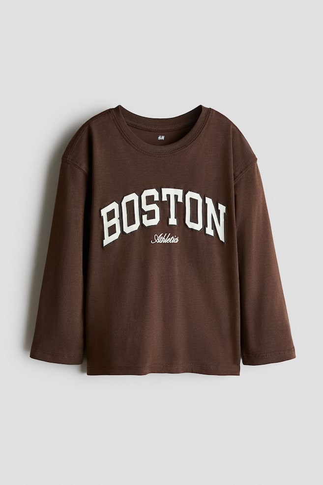 A brown long-sleeved t-shirt is displaying its front, featuring a ribbed crew neck. The shirt is showing "BOSTON" in large, white, raised appliqu� letters across the chest, with "Athletics" in smaller, white, embroidered script text positioned below. - 1