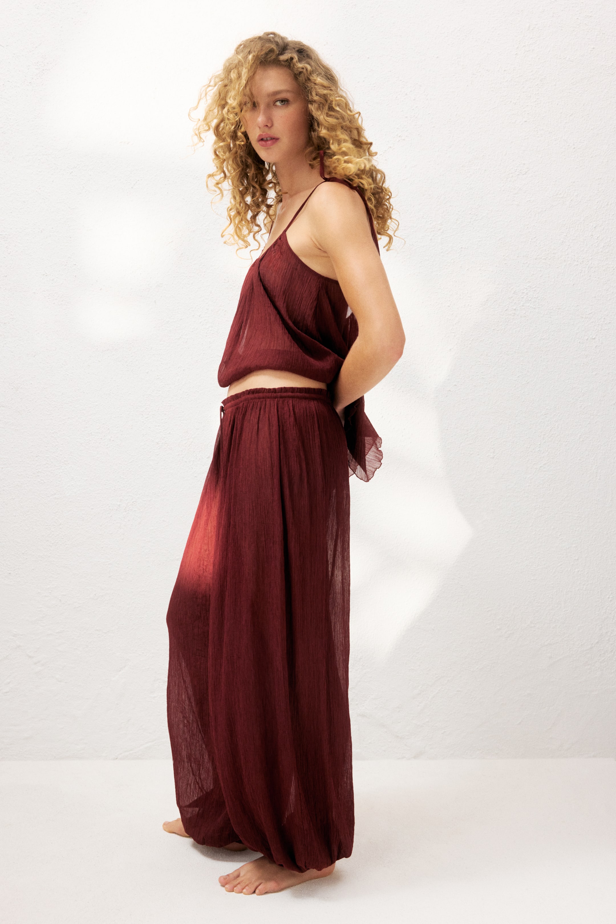 Ver imagen más grande: La modelo lleva unos pantalones de playa de corte holgado en un tejido transparente y arrugado de color rojo oscuro. Los pantalones cuentan con una cintura elástica y piernas abombadas con estrecha elasticidad en los dobladillos, combinados con un top a juego.