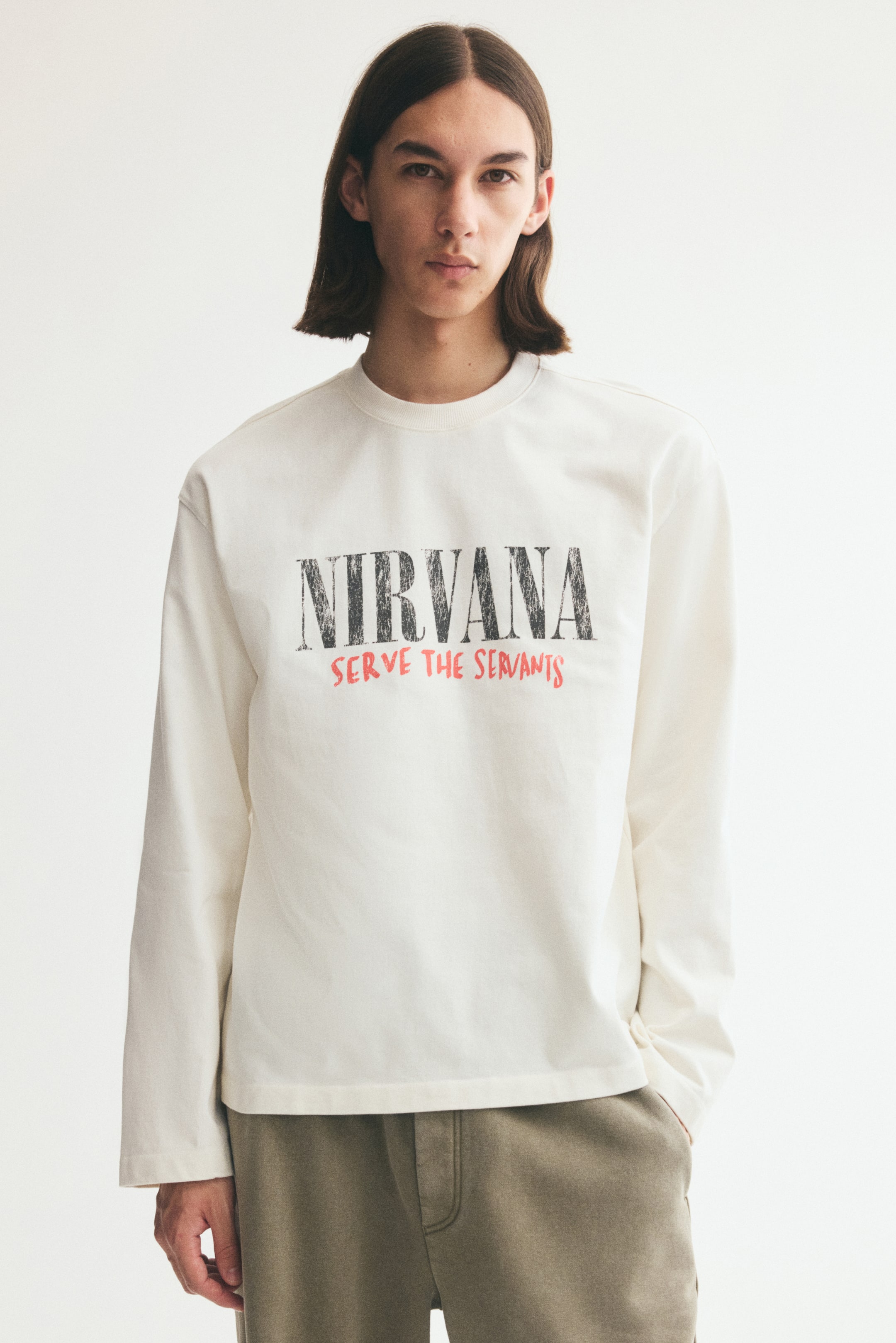 View larger image: A long-sleeve off-white cotton jersey tee is worn by the model, featuring a relaxed fit, dropped shoulders, and a ribbed round neckline. The front of the tee features a printed graphic with the word "NIRVANA" in distressed black capital letters above the phrase "SERVE THE SERVANTS" in red.