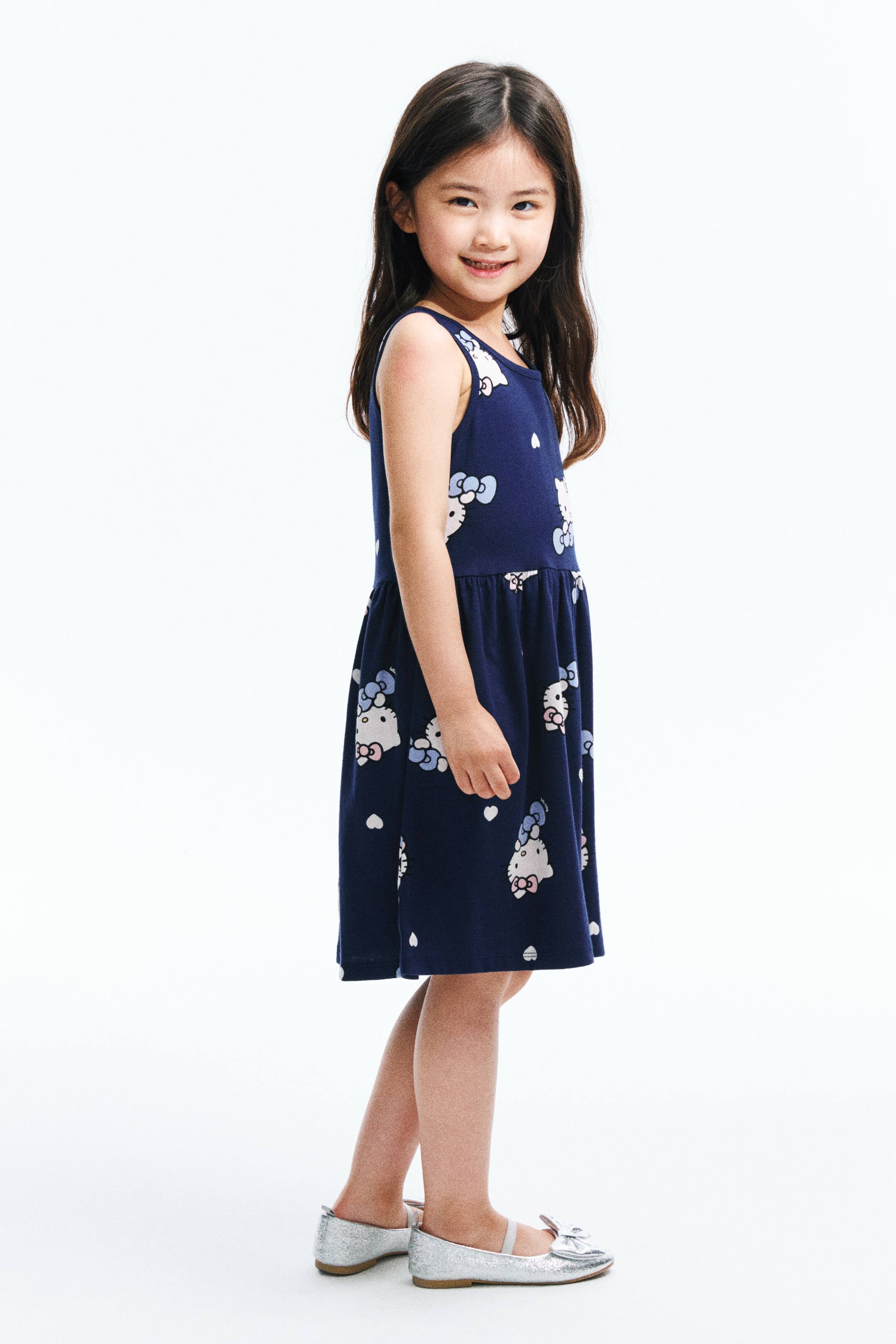 View larger image: "A navy blue sleeveless dress is showcased from a side angle. It highlights a gathered waist and a repeating pattern of white Hello Kitty characters with pink and blue bows, along with small white hearts scattered across its fabric."