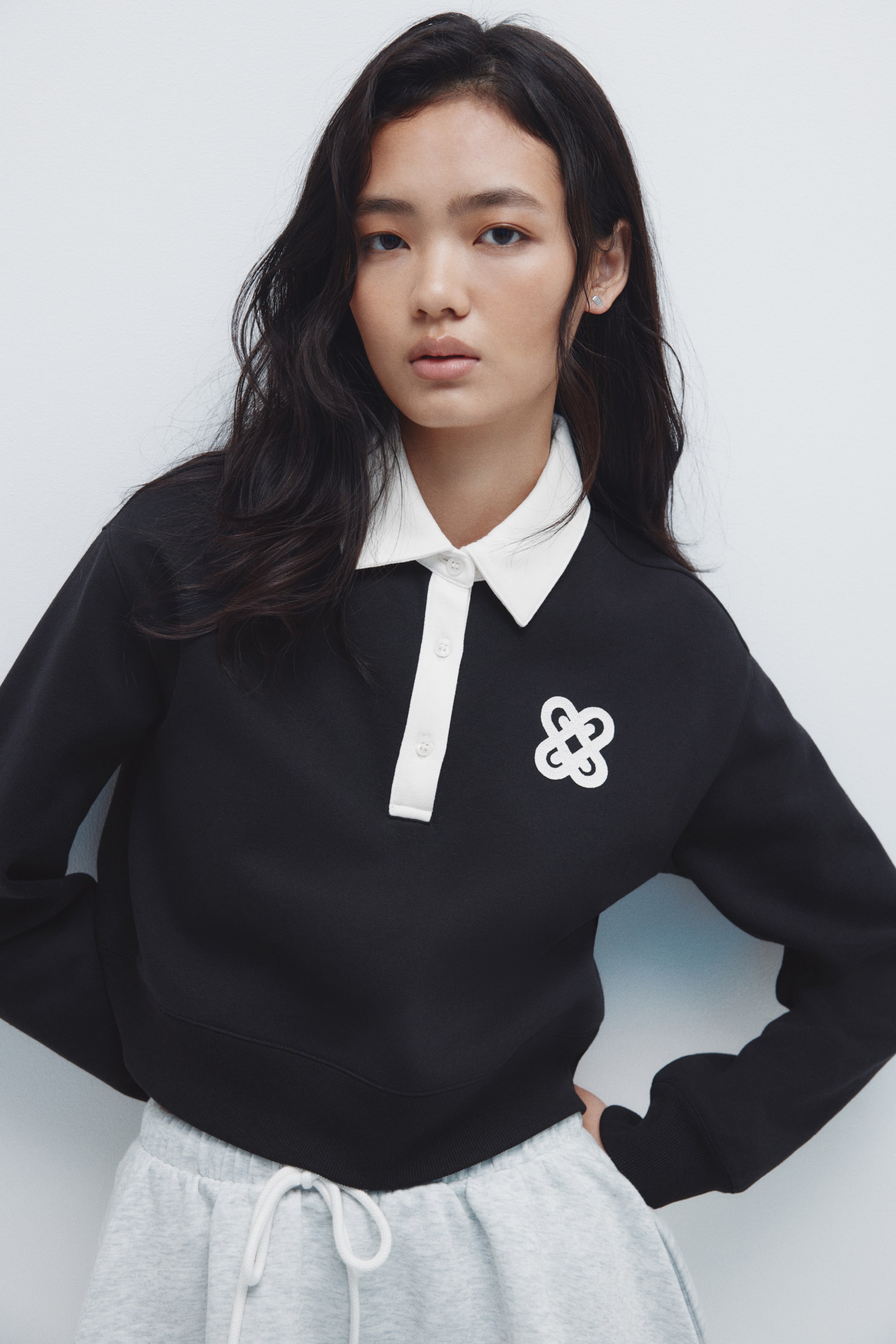 Xem ảnh lớn hơn: Một chiếc áo polo tay dài màu đen đang được mặc, nghiêng nhẹ sang bên trái, với cổ áo nhọn màu trắng và hàng nút ba màu trắng. Chiếc áo có hình logo trừu tượng màu trắng trên ngực trái và được thiết kế với cổ tay áo có gân và đường viền gân cropped, để lộ phần trên của chiếc quần thể thao màu xám.
