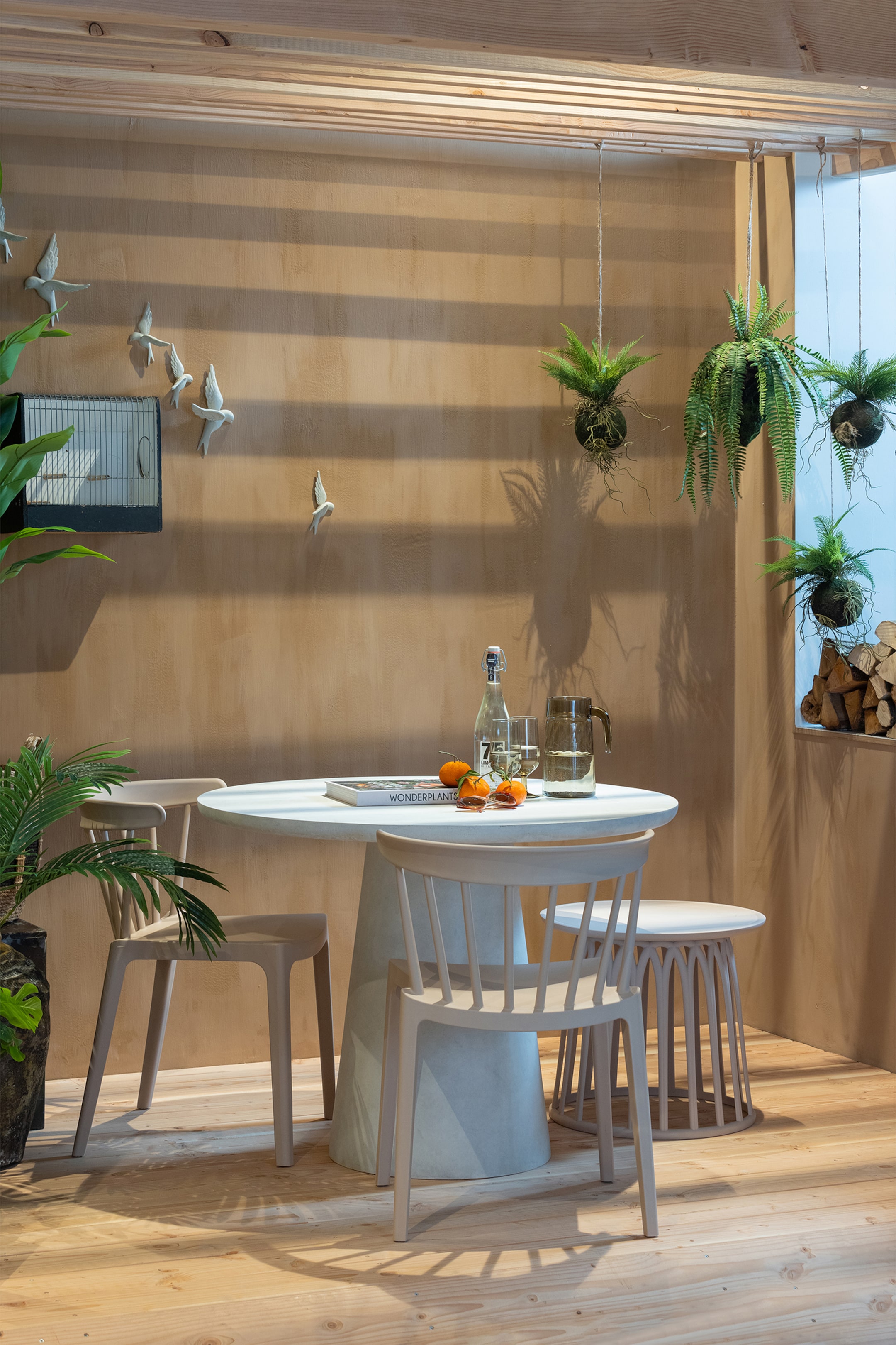 Grotere afbeelding bekijken: Een lichtgrijze ronde tafel, met een brede taps toelopende voet, staat in het midden. Twee bijpassende lichtgrijze stoelen, elk met een ronde zitting en een spijlenrugleuning, zijn rond de tafel geplaatst, waarbij één stoel naar voren is gericht en de andere iets naar links is gedraaid. Een bijpassende lichtgrijze kruk, met een ronde zitting en een gegroefde voet, is ook zichtbaar naast de tafel.