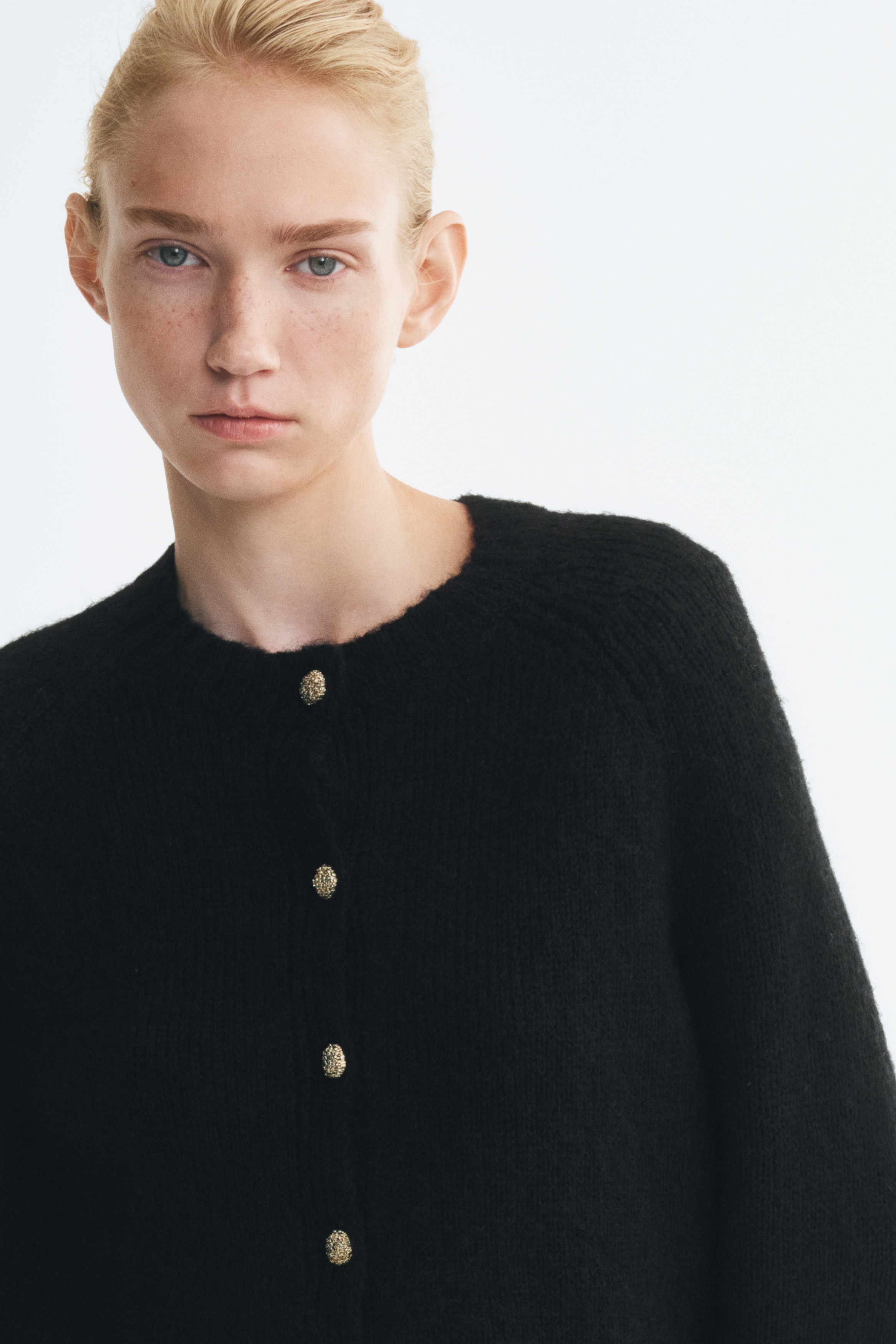A black knit cardigan is shown from the front, featuring a soft, fuzzy texture, a round neckline, and long raglan sleeves with ribbed cuffs. It features a front button closure with six textured, gold-tone buttons and a ribbed hem.