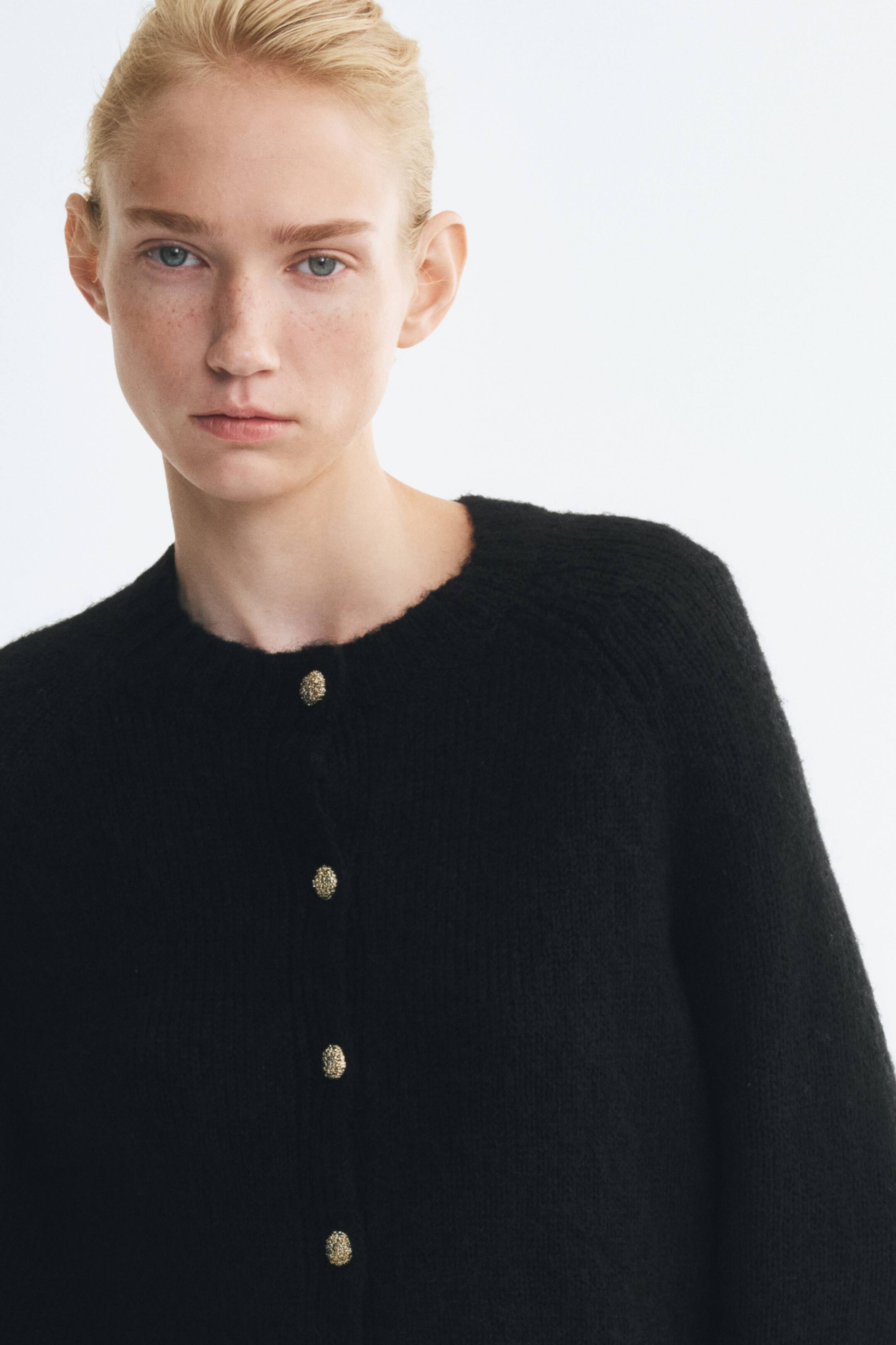 View larger image: The model is wearing a black soft-knit cardigan with textured gold-tone buttons. The cardigan features a round neckline with rib trim and raglan sleeves.