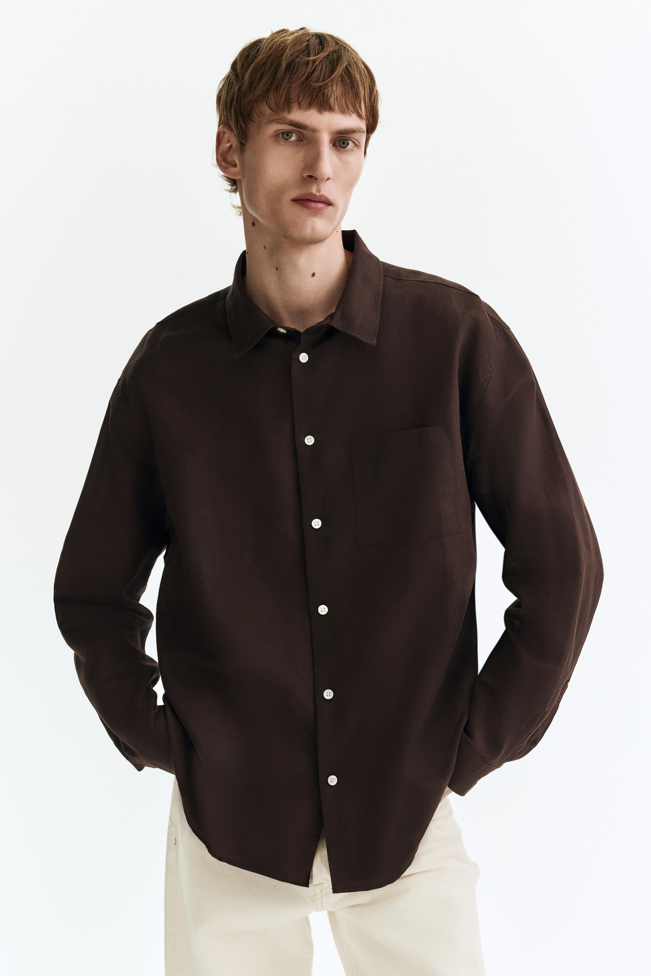 View larger image: A dark brown long-sleeved shirt features a visibly textured fabric, a pointed collar, and a button-front closure with white buttons. A patch pocket is positioned on the left chest, and a curved hem shapes the bottom.