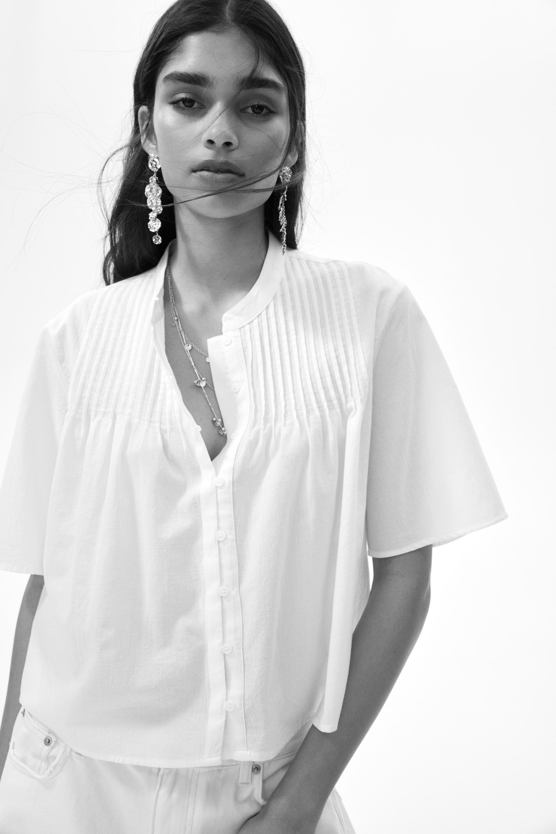 View larger image: A white shirt featuring a band collar and vertical pin-tucks on the chest, extending from the shoulder line. It showcases a partial button-down placket with visible buttons, wide short sleeves, and a silver necklace layering over the partially open neckline.