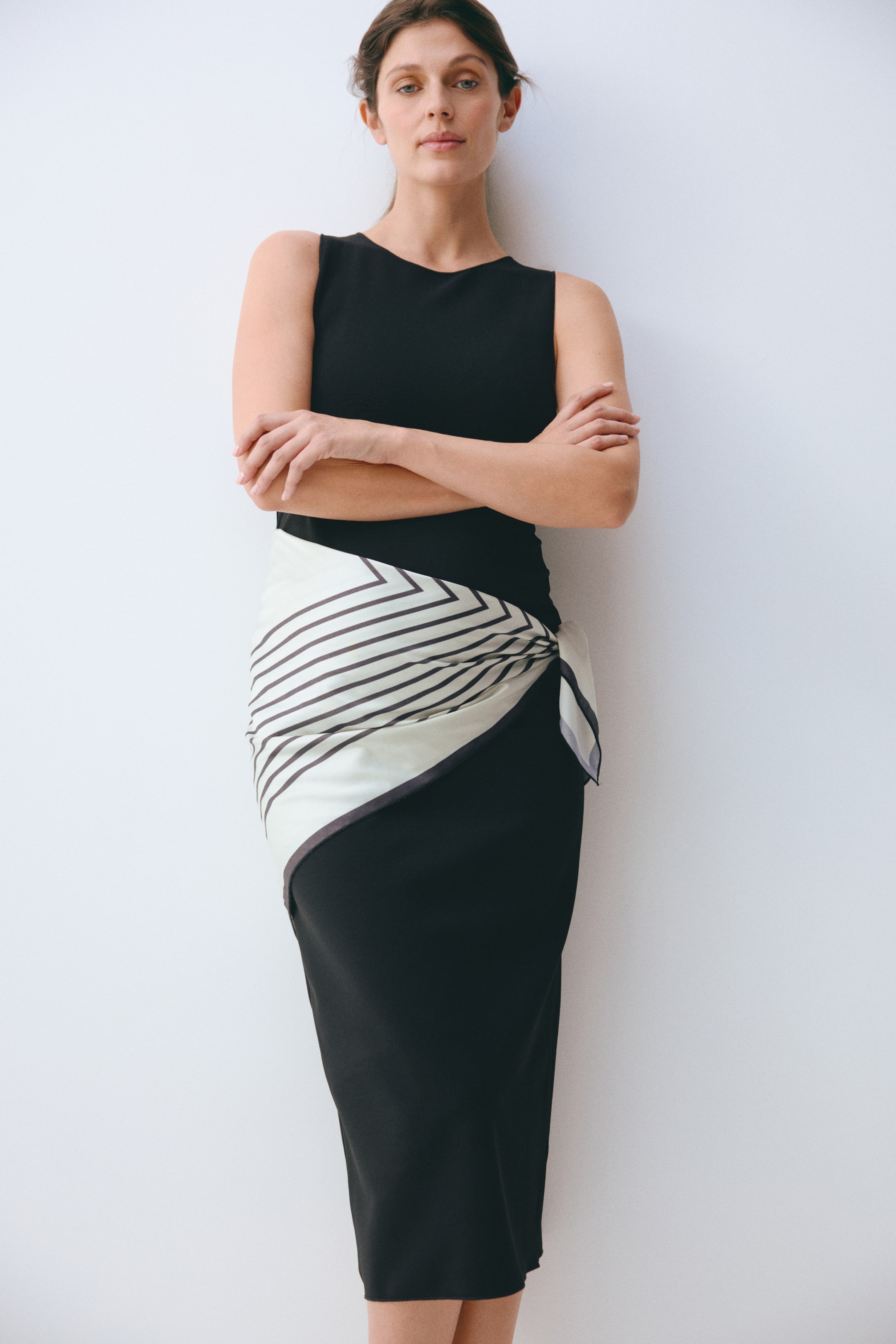 Agrandir l'image: Un haut noir sans manches avec un col rond est porté avec une jupe crayon noire qui descend sous le genou. Un foulard couleur crème avec des rayures diagonales foncées formant un motif chevron est noué autour de la taille, couvrant la partie supérieure de la jupe.