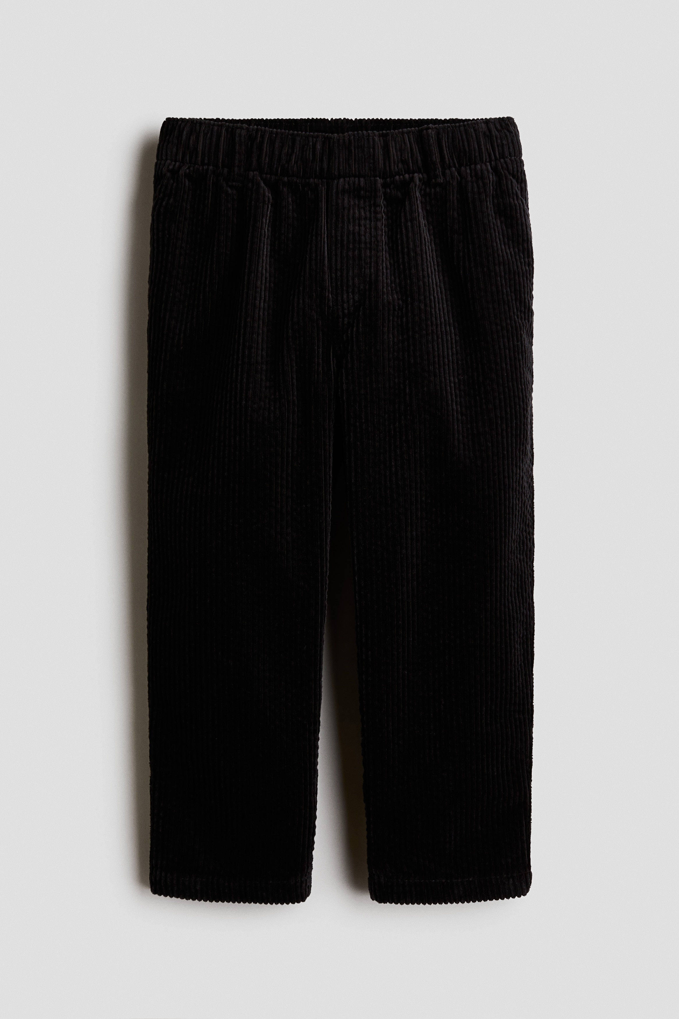 Ce pantalon en velours cÃ´telÃ© brun foncÃ© H&M Adorables est prÃ©sentÃ© de face et se distingue par sa coupe droite et sa texture cÃ´telÃ©e bien visible. Le pantalon est agencÃ© avec une chemise boutonnÃ©e Ã  carreaux rouges et des chaussures plates noires, tandis quâune main repose dans une poche latÃ©rale.