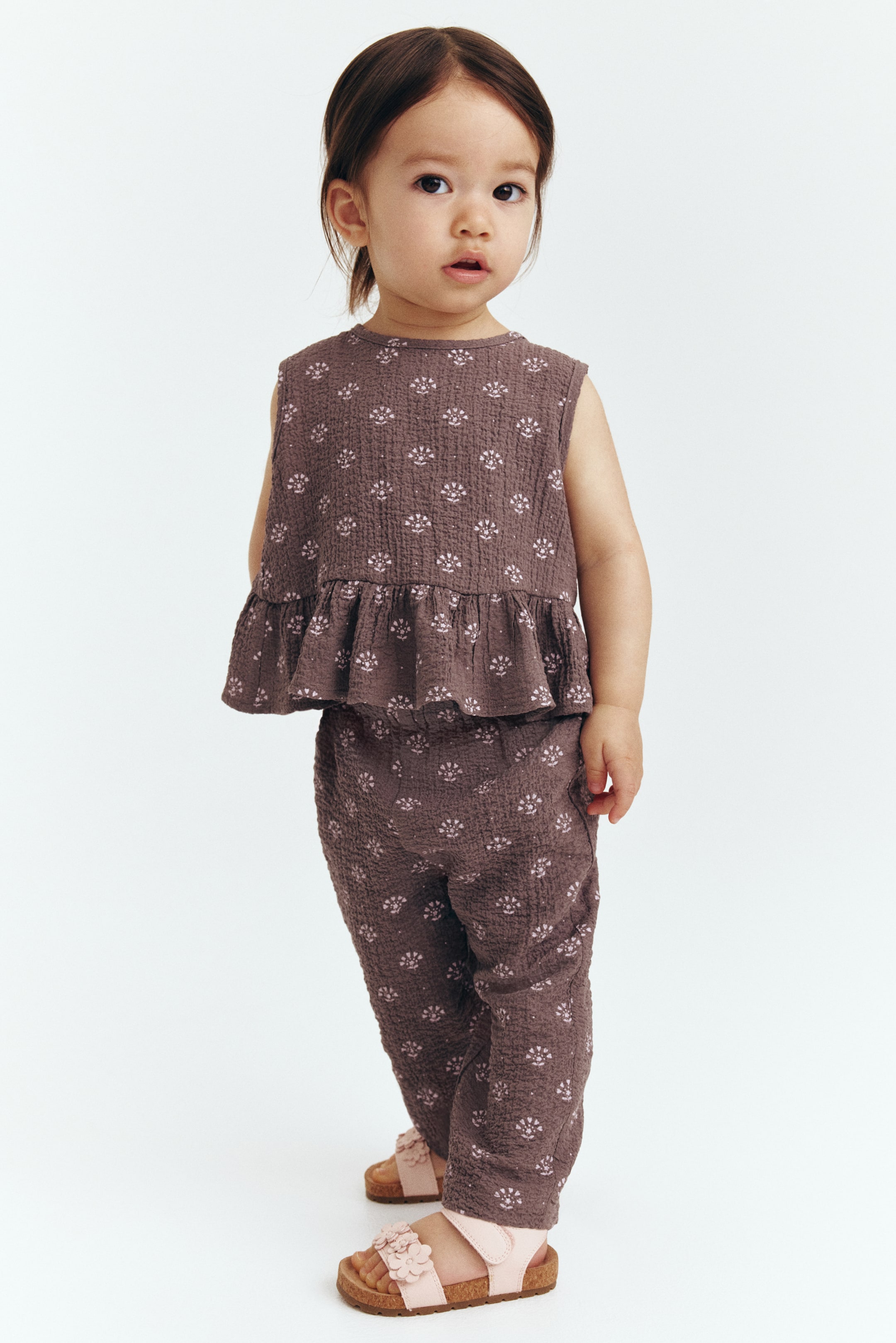 View larger image: A two-piece children's outfit is displayed at a slight angle, showcasing a dusty mauve textured fabric adorned with light pink floral motifs. The sleeveless top features a round neckline and a ruffled peplum hem, while the matching full-length pants offer a comfortable loose fit.