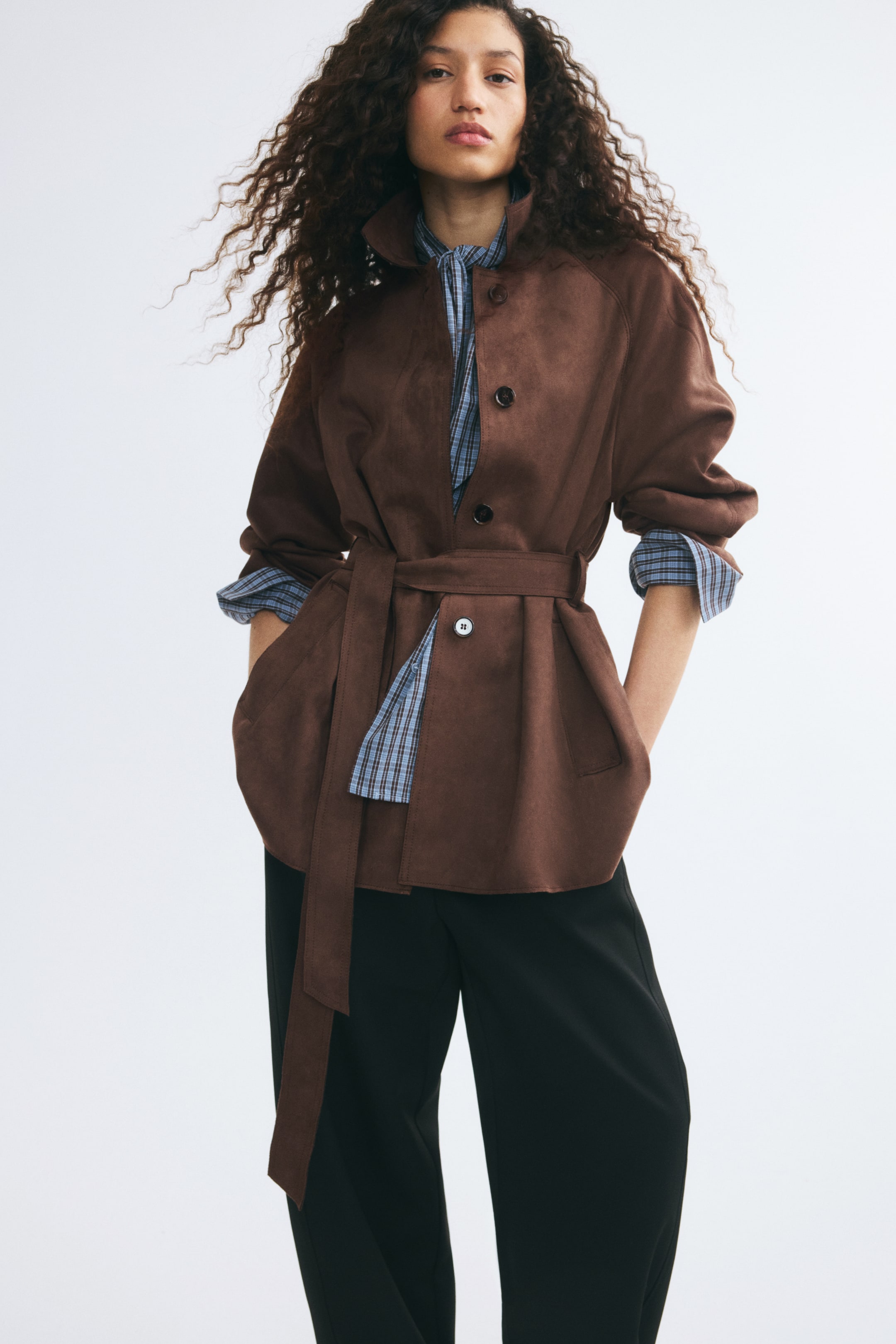Agrandir l'image: Le modèle porte un court manteau brun à la finition douce et légèrement lainée, doté d’un col montant, d’une fermeture boutonnée sur le devant et d’une large ceinture assortie nouée à la taille. Le manteau est porté sur une chemise à carreaux bleus et un pantalon noir, tandis que le modèle repose ses mains dans les poches passepoilées avant.
