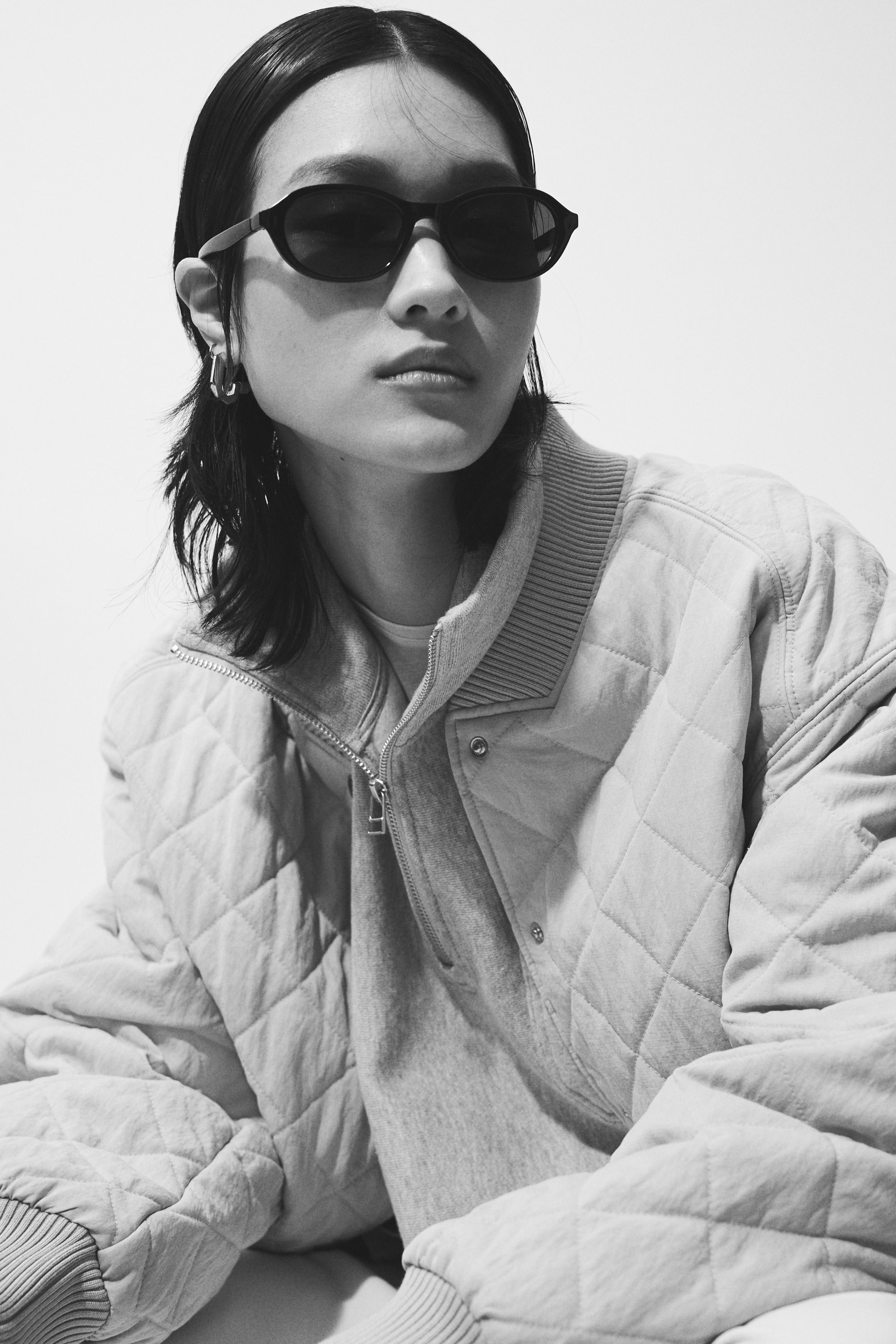 Προβολή μεγαλύτερης εικόνας: Ένα ανοιχτόχρωμο καπιτονέ μπουφάν παρουσιάζει ένα σχέδιο καπιτονέ με διαμάντια, ένα ριμπ όρθιο γιακά και μπροστινό κλείσιμο με κουμπιά τύπου σούστα. Φοριέται ανοιχτό, πάνω από ένα γκρι φούτερ με φερμουάρ, και οι ριμπ μανσέτες του είναι ορατές.