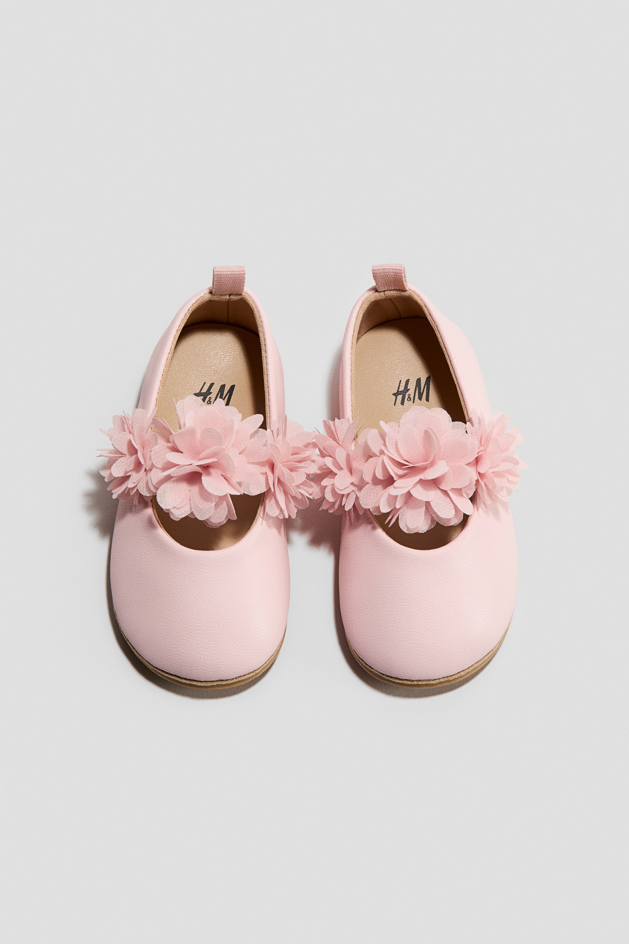 View larger image: Two pink baby shoes are displaying a front view, positioned side by side. Each shoe is featuring a rounded toe, a light brown sole, a small fabric loop at the heel, and a cluster of layered pink fabric flowers adorning its front upper, with the H&M logo showing on the insole.