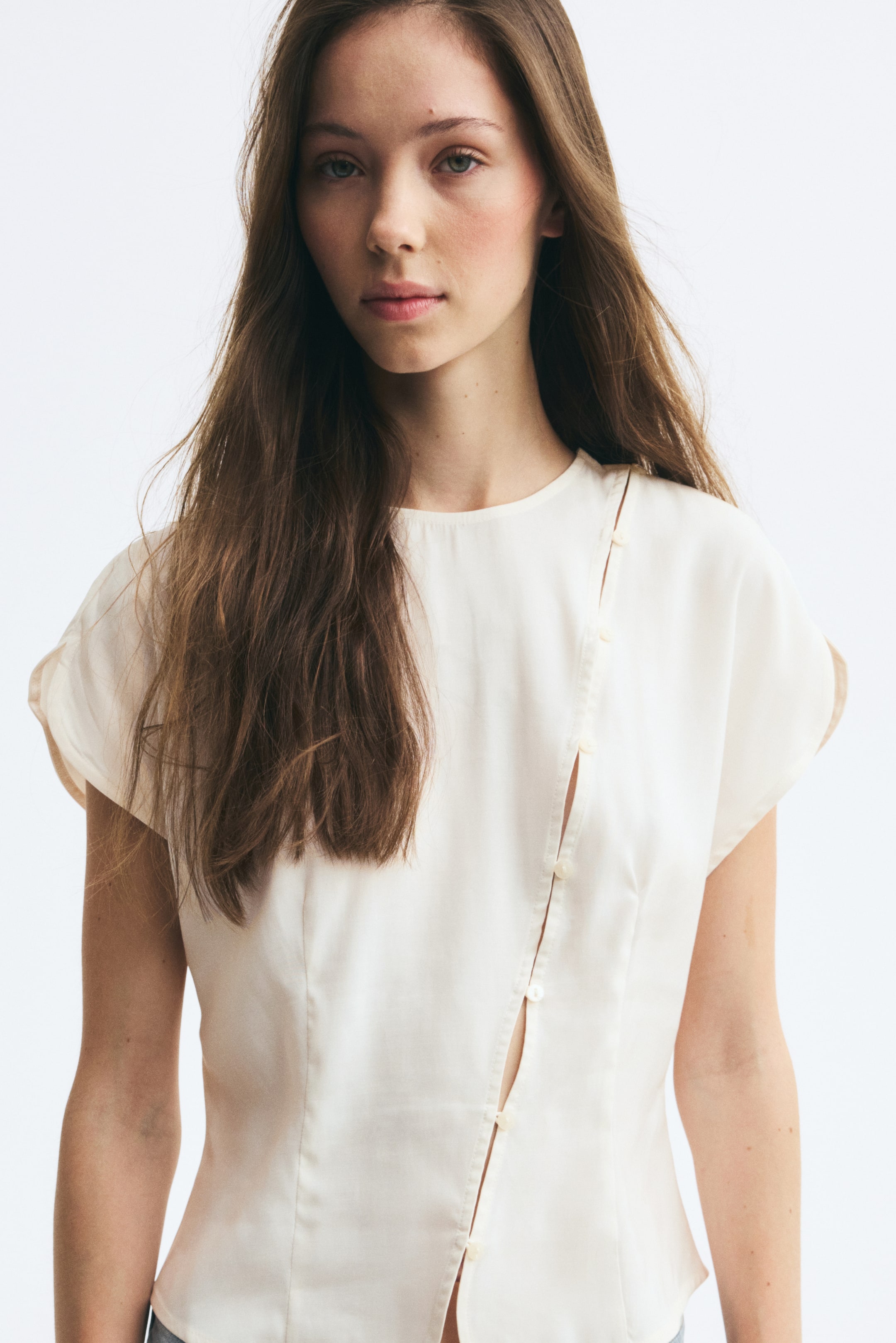 View larger image: The model is wearing a cream-colored blouse featuring a round neckline, cap sleeves, and shaping waist darts. The blouse is designed with a diagonal row of button-and-loop fasteners running from one shoulder down the front.