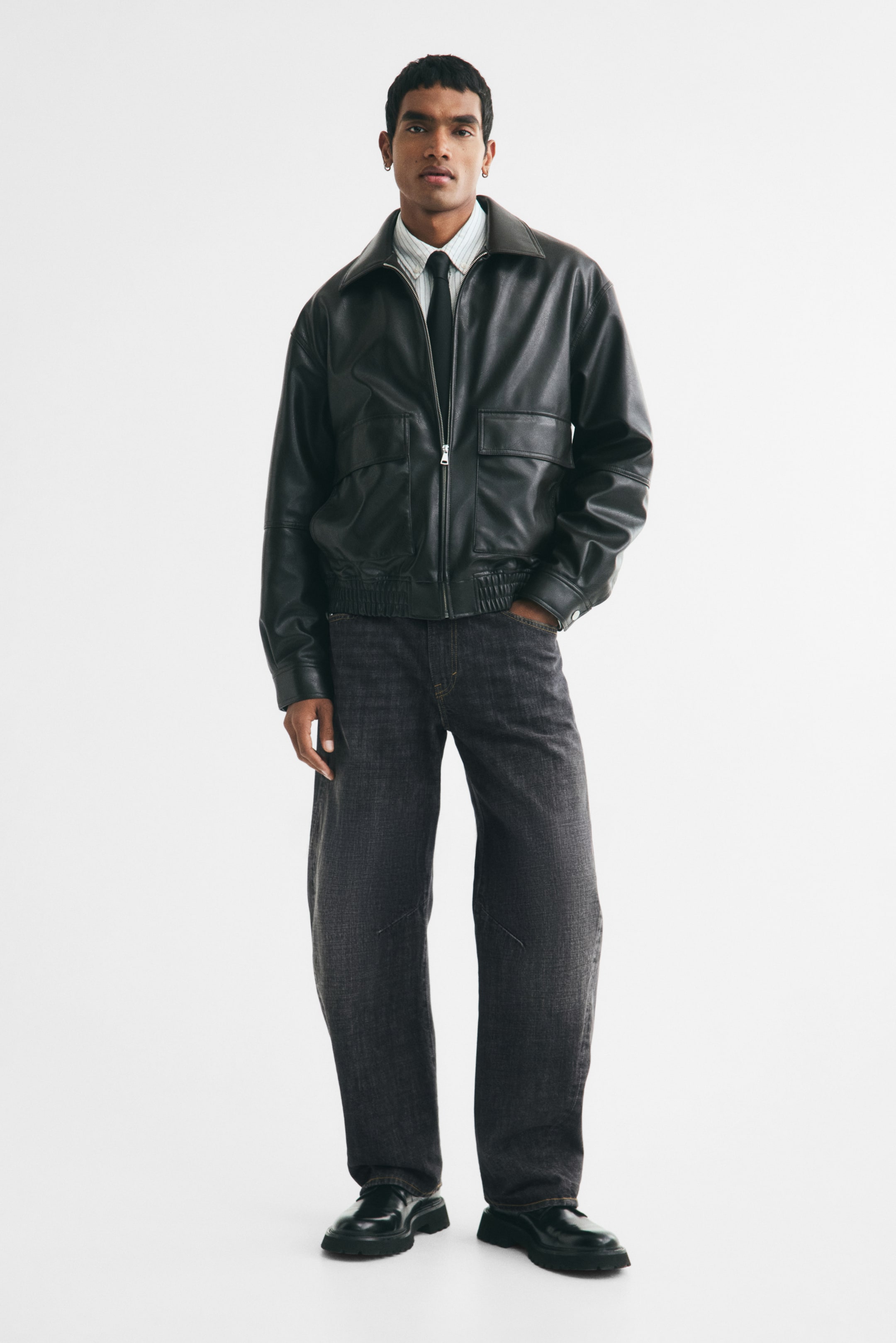 View larger image: The model is wearing a black faux leather bomber jacket over a striped shirt and black tie. The jacket features a pointed collar, a front zip closure, and two flap pockets.