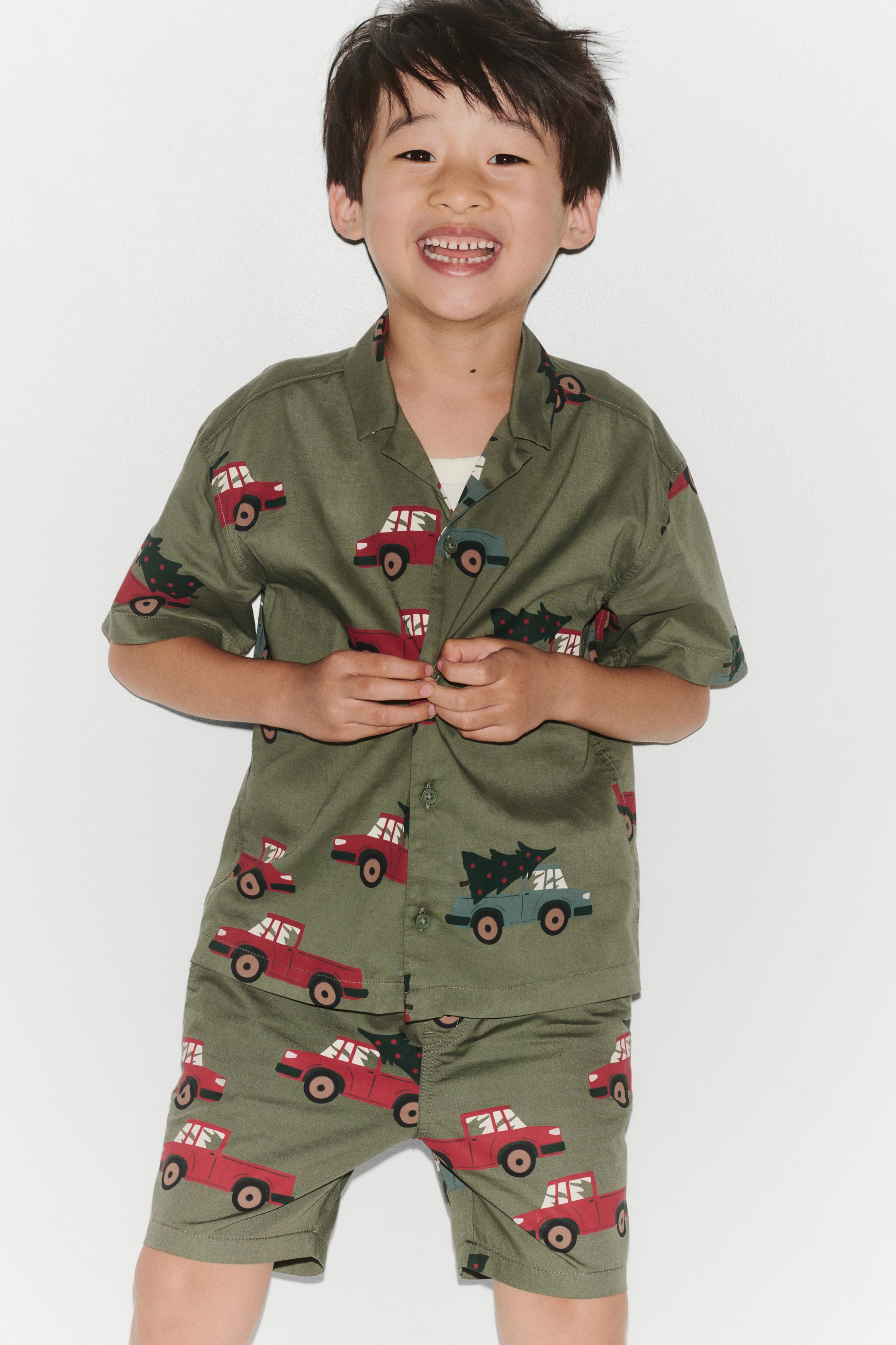 View larger image: A green, short-sleeve shirt featuring an all-over print of red and blue pickup trucks carrying pine trees is shown from the front. The shirt features a resort collar, front button closure, and gently dropped shoulders, and is styled with matching printed shorts.