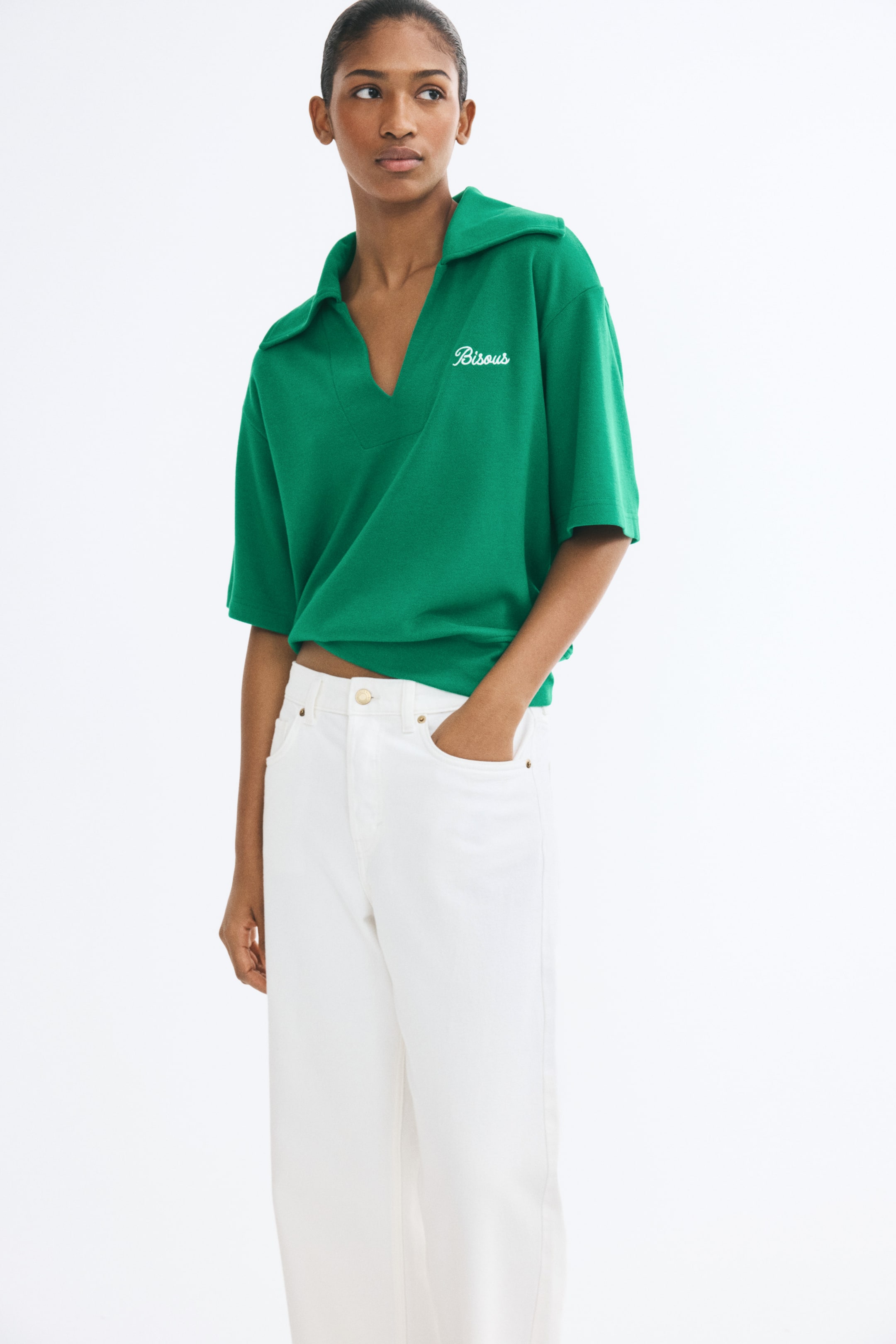 Ampliar la imagen: La modelo lleva puesta una playera tipo polo verde de manga corta, con cuello abierto amplio de doble capa y texto bordado en blanco en el pecho. La prenda tiene hombros caídos y una pretina ajustada que crea una silueta ligeramente abombada, y se combina con jeans blancos.