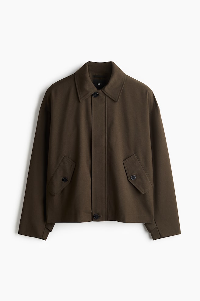 Eine dunkel olivgrüne Jacke wird von vorne gezeigt, mit einem Umlegekragen, einer verdeckten Knopfleiste mit sichtbaren oberen und unteren Knöpfen, zwei schrägen aufgesetzten Taschen mit geknöpften Klappen an der unteren Vorderseite, langen Ärmeln und einem geraden Saum. - 1