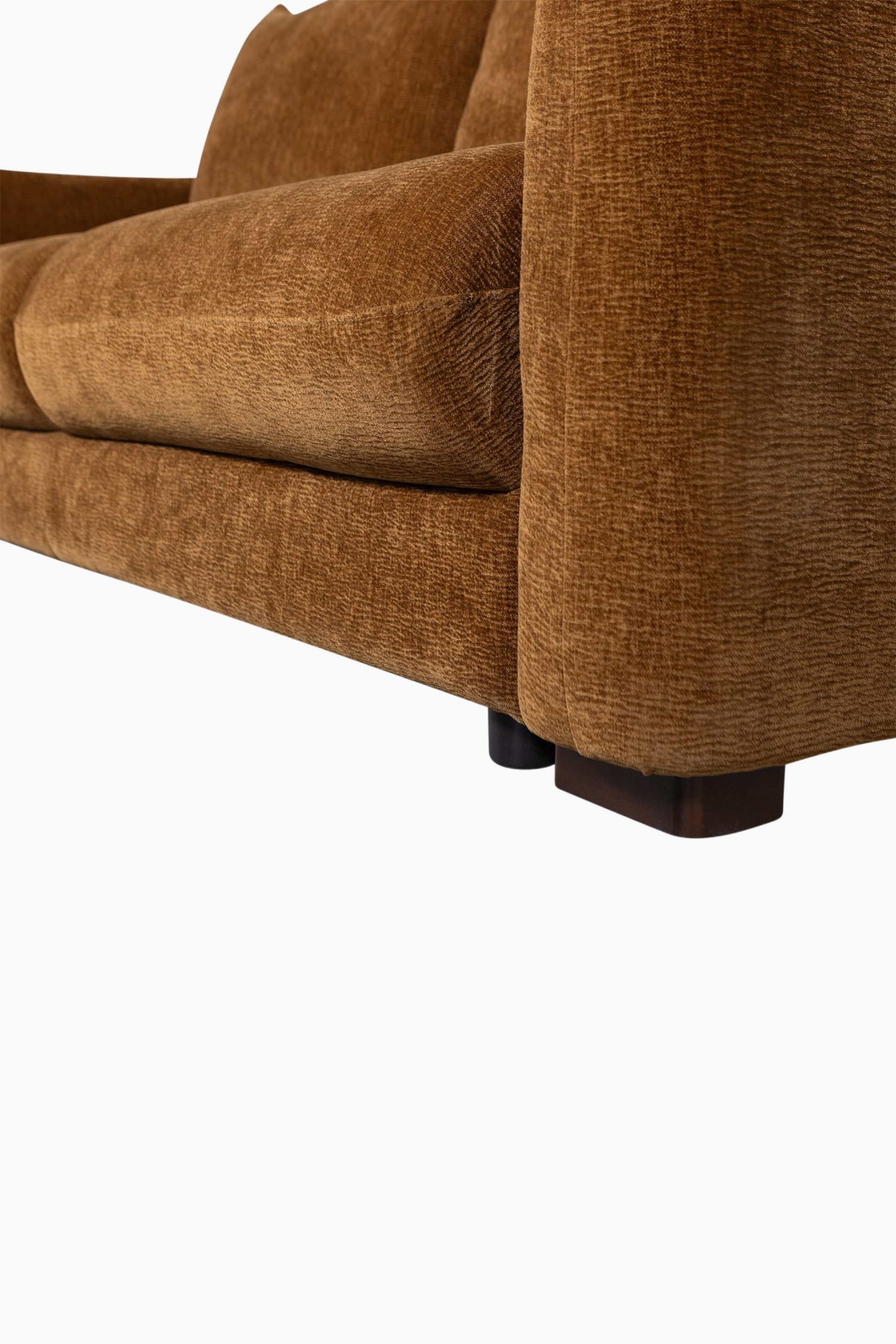 Grotere afbeelding bekijken: Een okerkleurige bruine sofa is vanuit een laag perspectief onder een hoek geplaatst, waardoor de voorzijde aan de rechterkant zichtbaar is. Het toont een gestructureerde bekleding die dikke zit- en rugkussens bedekt, een brede armleuning, en donkere blokpoten die de basis ondersteunen.