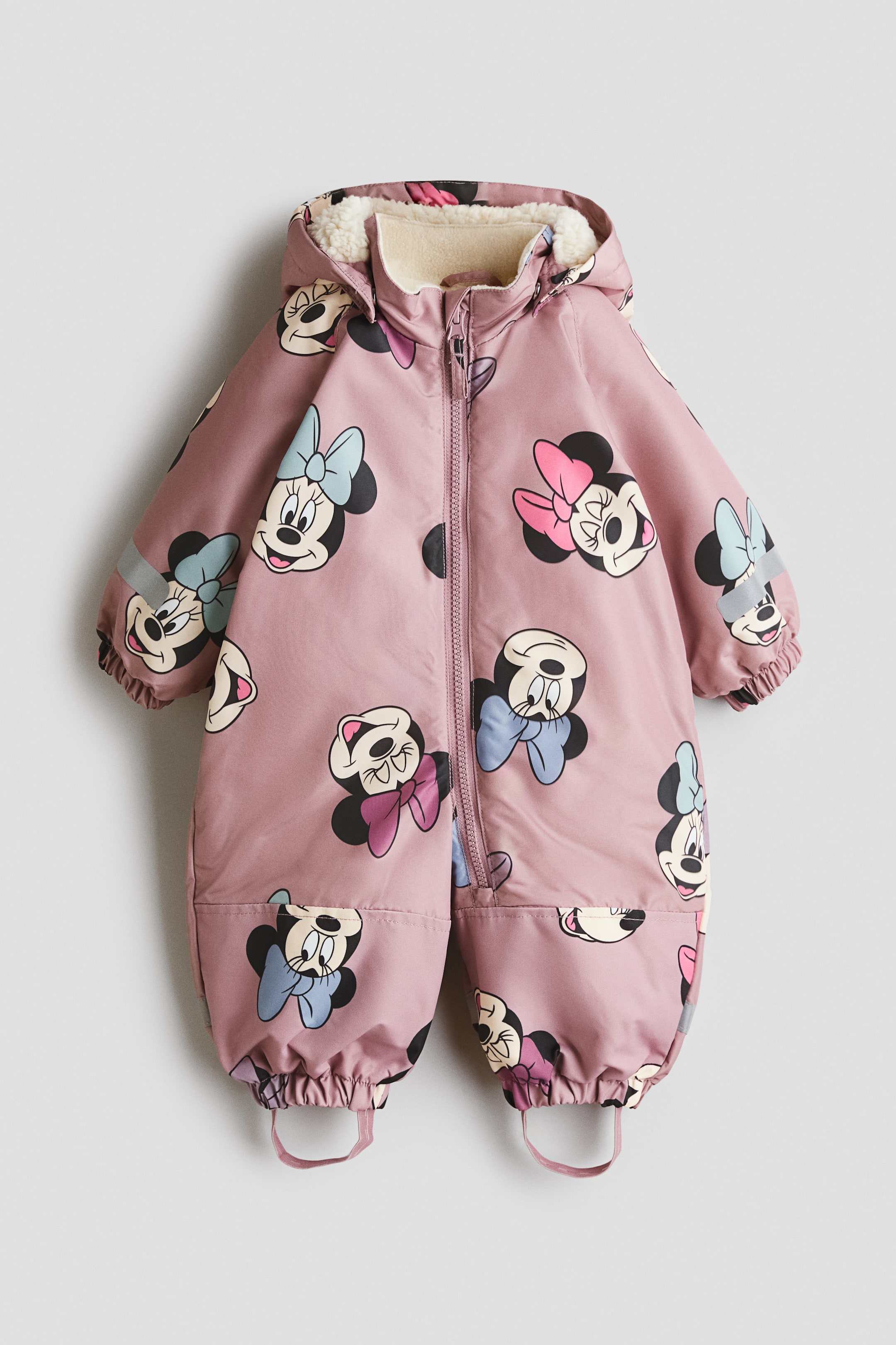 View larger image: A pink baby snowsuit features an all-over Minnie Mouse print with multicolored bows. It is designed with a central front zipper closure and an attached hood lined with cream sherpa, and includes elasticized cuffs, with one sleeve featuring a reflective strip, as well as elasticized ankles with stirrups.