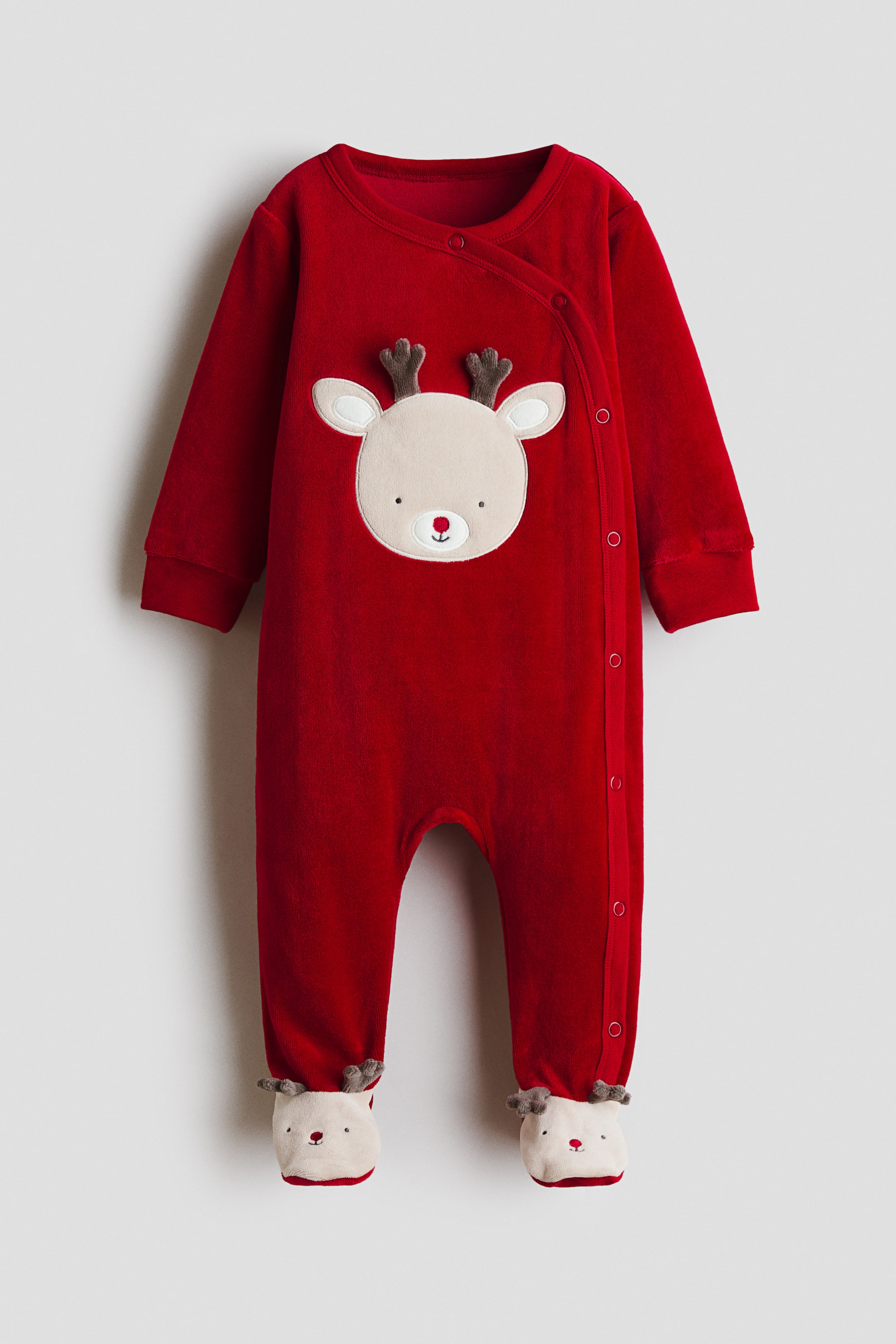Agrandir l'image: Une combinaison de nuit rouge pour bébé arbore une applique de visage de renne de couleur claire sur la poitrine, avec des bois marron, des oreilles blanches, des yeux noirs et un nez rouge. Elle présente une fermeture asymétrique à pressions le long du côté droit de l’avant et se termine par des pieds intégrés conçus comme des têtes de renne assorties.