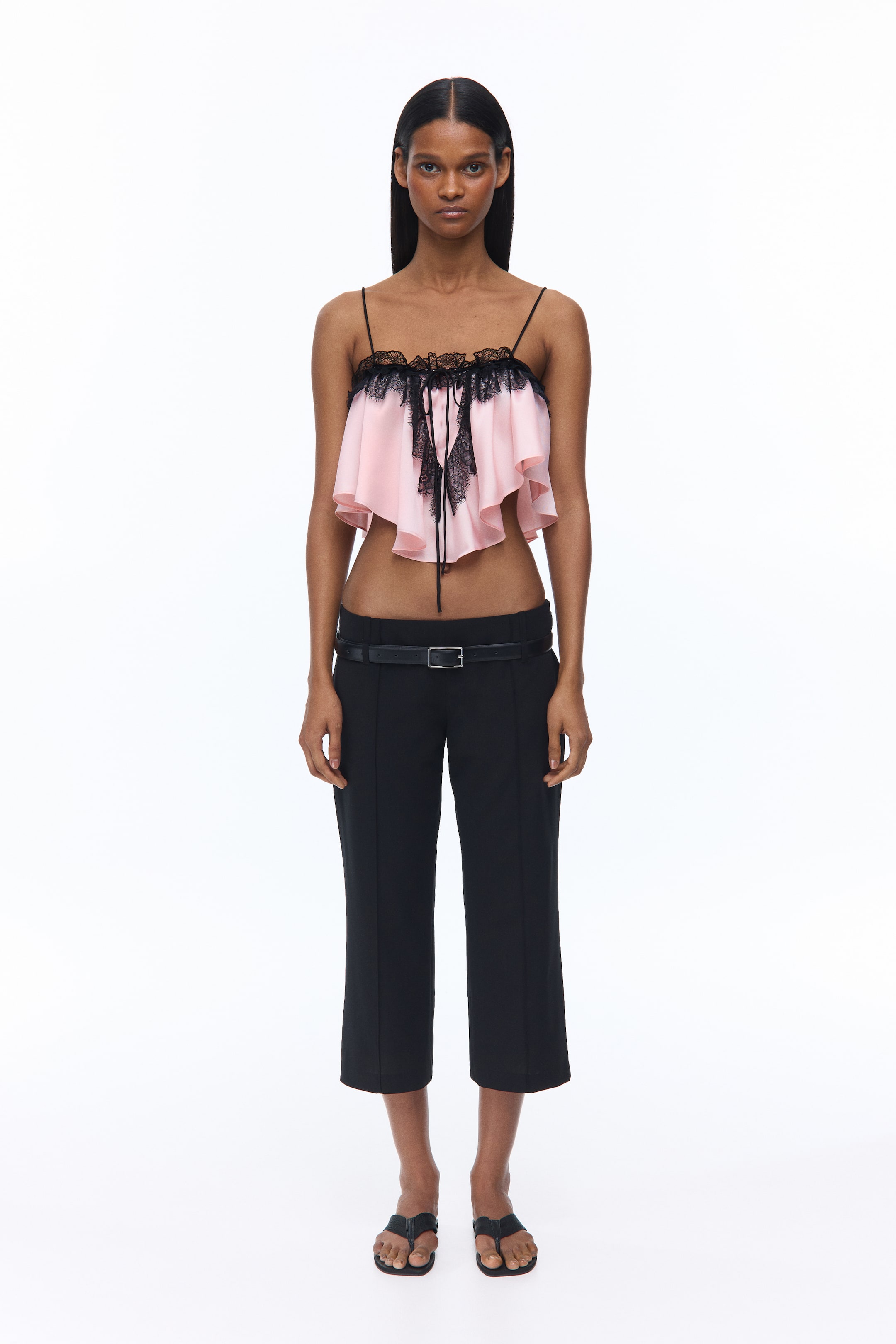 View larger image: The model is wearing a pink loose-fit satin crop top with a straight neckline accented by black scalloped lace ruffles. It features an interwoven black satin spaghetti tie fastened at the front. A wider black lace ruffle, adorned with an eyelash-stitch trim, runs parallel to the neckline ruffles and transitions into a V-shape at the front. Black spaghetti shoulder straps thread through small loops, and the top finishes with a rounded hem.