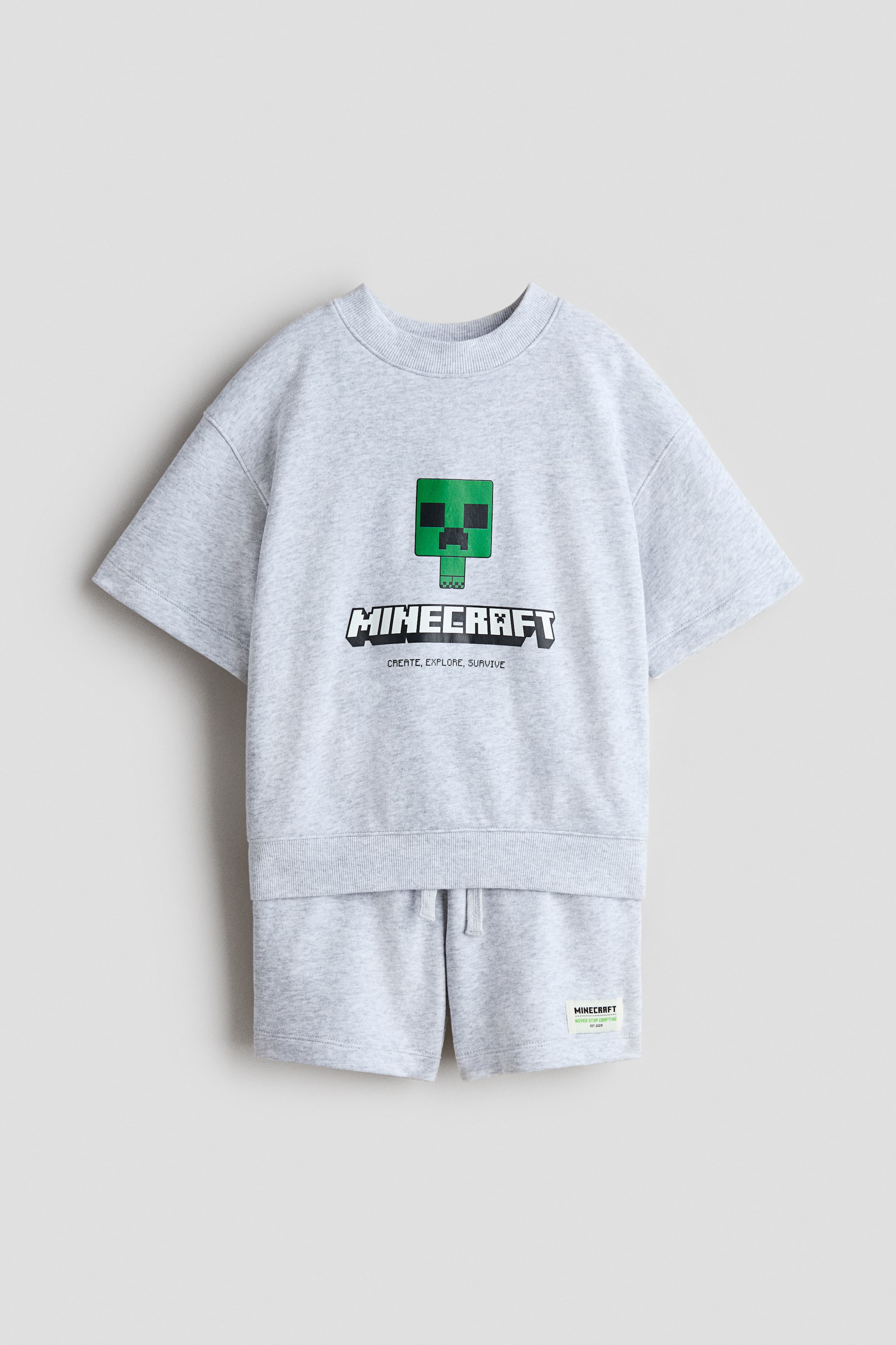 הצגת תמונה גדולה יותר: סט החולצה והמכנסיים בצבע אפור בהיר מוצג מלפנים, עשוי מבד סווטשירט קליל. החולצה מציגה דמות פיקסלית ירוקה ולוגו "MINECRAFT" על החזה, עם צווארון עגול מצולע, כתפיים מורדות, שרוולים קצרים ורחבים ושוליים מצולעים, בעוד שהמכנסיים מציגים מותן אלסטית עם שרוך ותווית קטנה של Minecraft על הרגל הימנית.