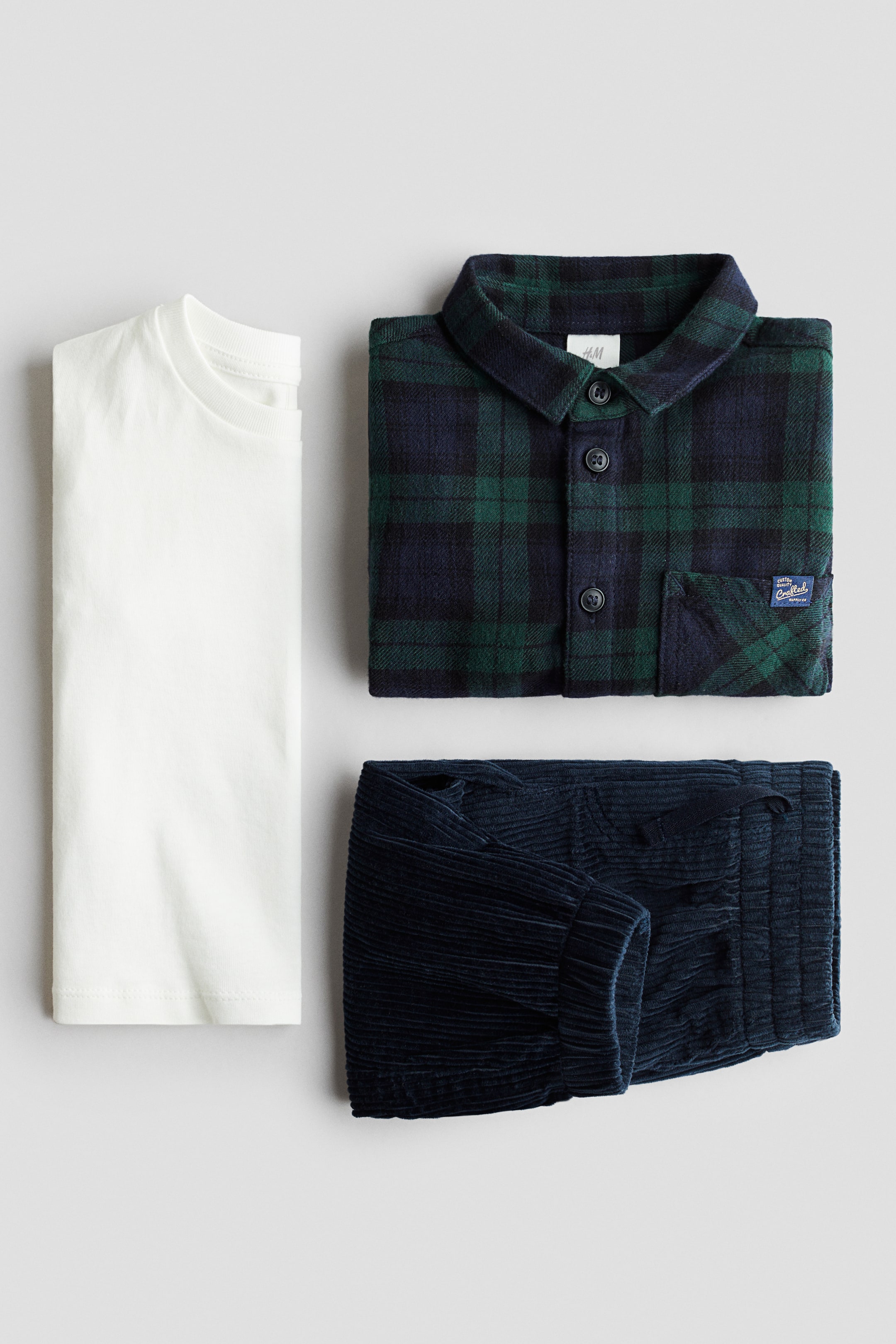 Voir une image plus grande: Un ensemble en coton de 3 pièces est exposé, comprenant un t-shirt blanc uni à col rond plié. Sont également présentés une chemise à carreaux verte et marine pliée, montrée de FACE avec un col classique et une poche poitrine, ainsi qu'un pantalon en velours côtelé bleu marine plié avec une taille élastiquée à cordon de serrage et des poignets élastiqués.