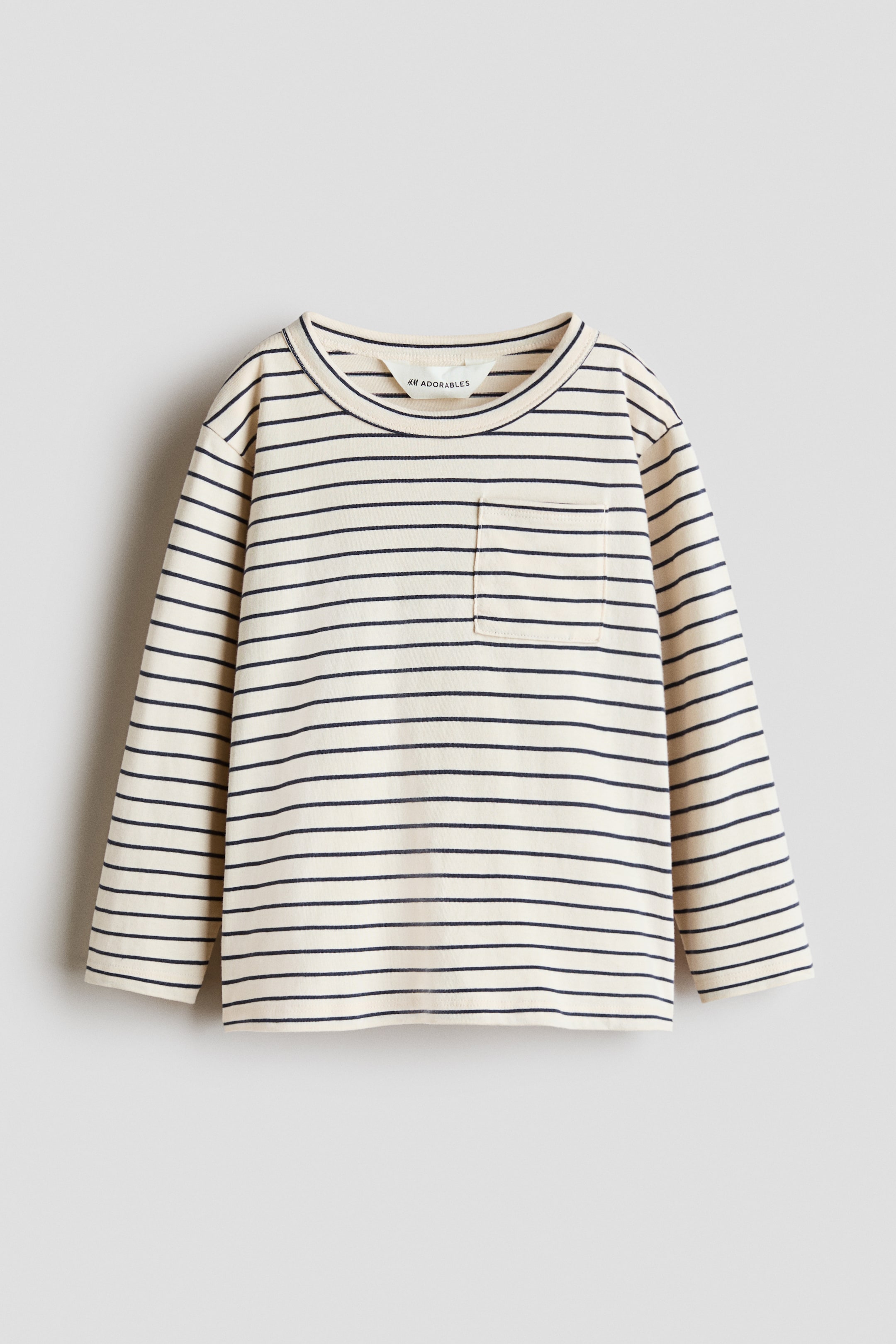 拡大画像を表示: 長袖Tシャツが平置きされ、正面を向いており、クリーム色の生地に細いネイビーの横ストライプが入っています。リブ仕様のクルーネックで、左胸にはパッチポケットが付いています。