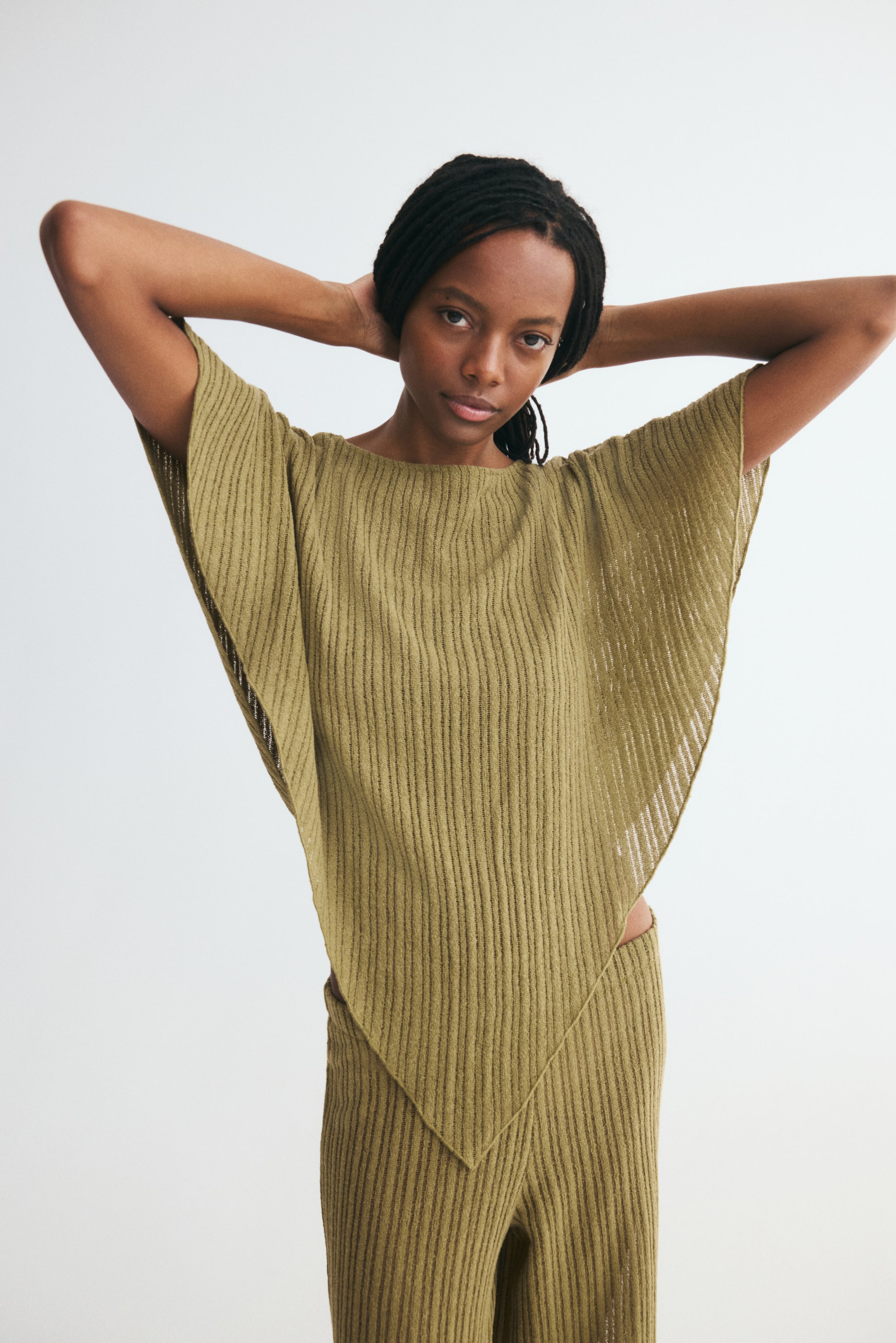 Visualizza l'immagine ingrandita: Un morbido mantello in maglia a coste verde oliva è mostrato frontalmente, con scollo tondo, ampi lati drappeggiati e orlo asimmetrico a fazzoletto. Il mantello è abbinato a pantaloni coordinati in maglia a coste, mettendone in risalto la silhouette ampia e fluida.