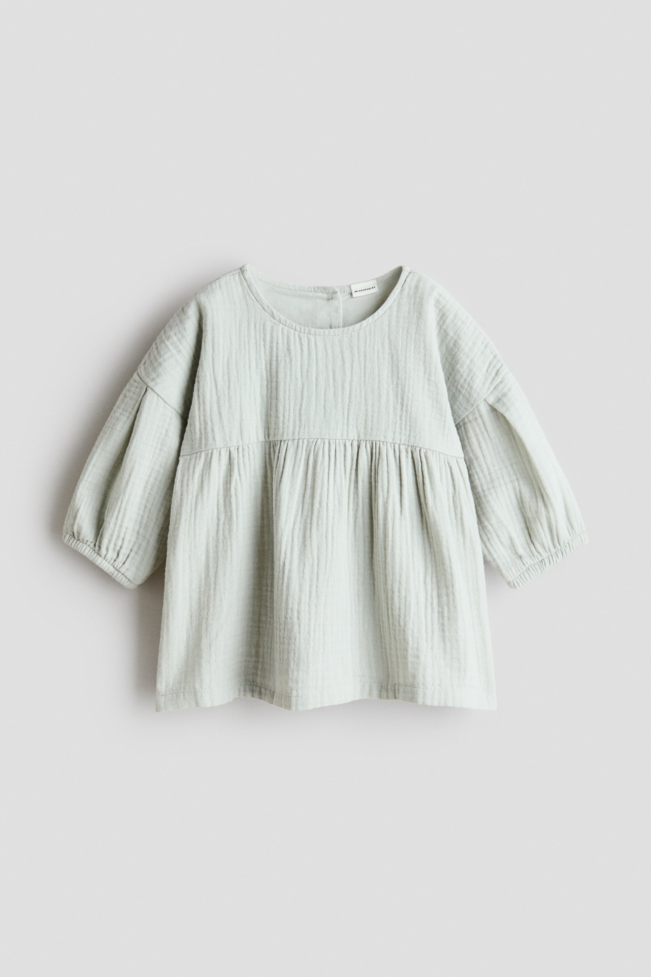 View larger image: An H&M Adorables dress is shown from the front, featuring a light green muslin fabric with a round neckline. It is designed with low dropped shoulders, long puffy sleeves with elasticated cuffs, and a gathered seam at the waist leading to a voluminous skirt.