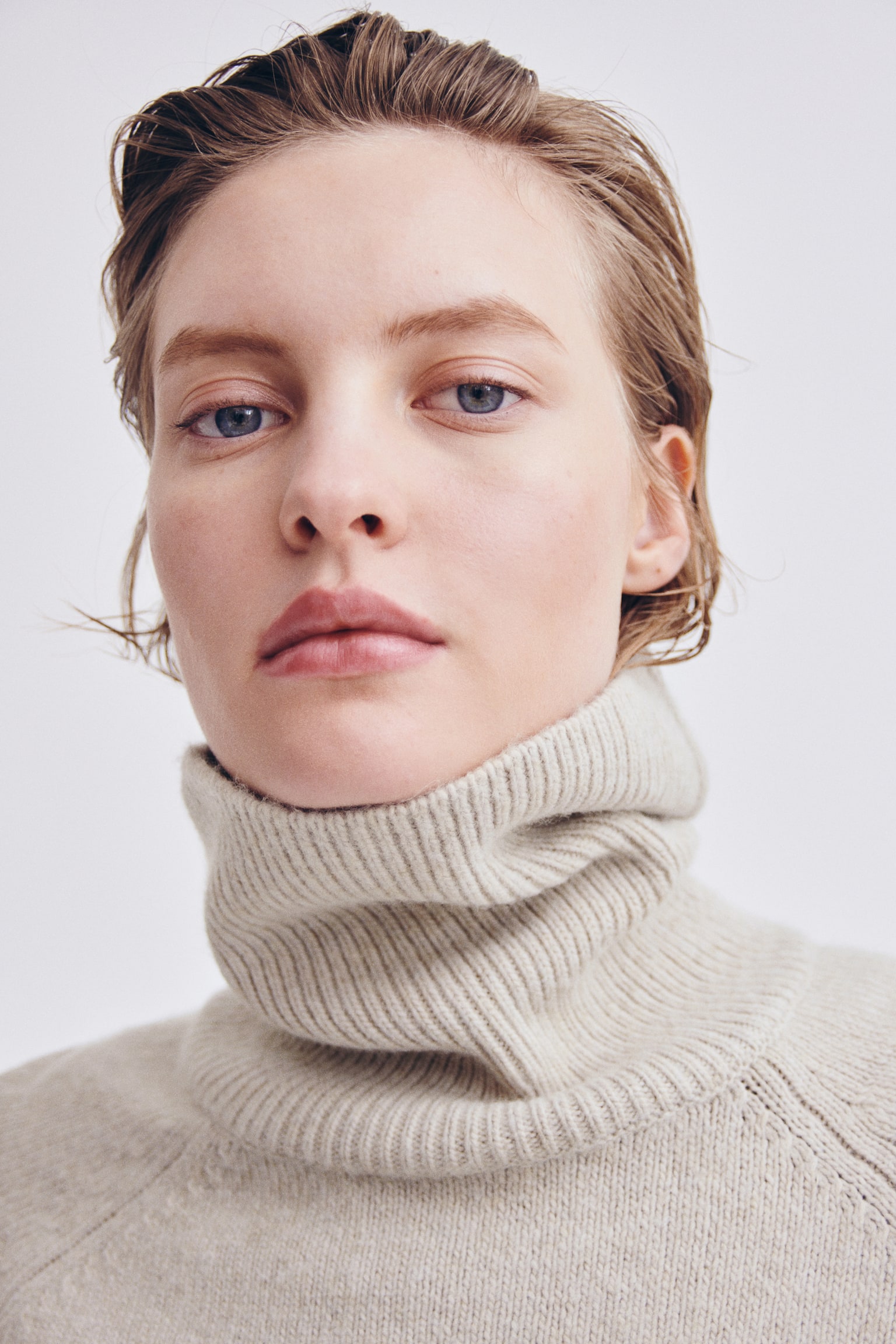 Un pull en maille douce beige clair, avec un col roulé côtelé montant et des coutures d’épaules raglan, est porté par le modèle dans un portrait en gros plan. - 1