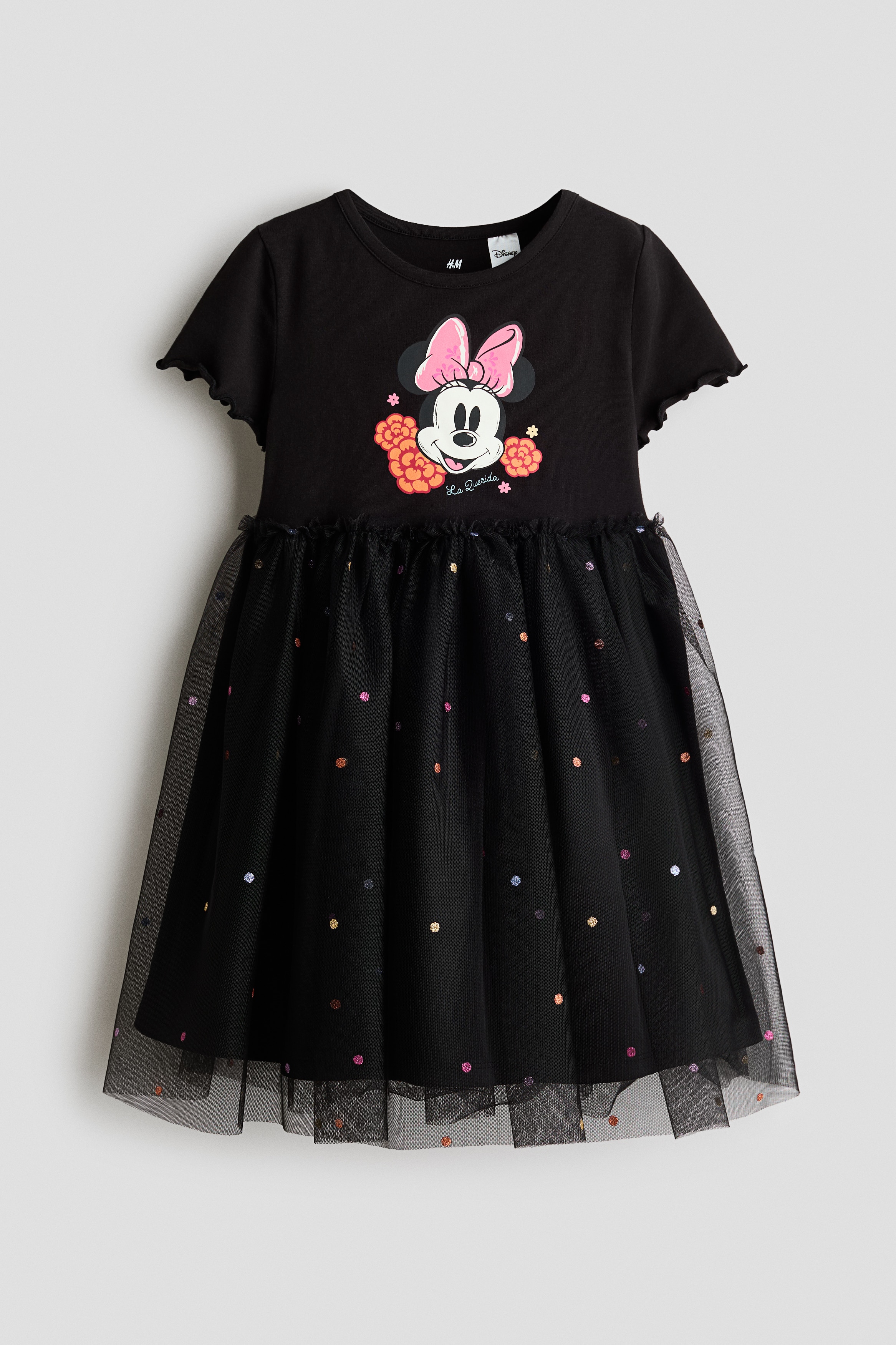 A child is wearing a black jersey dress, shown from the front, featuring a Minnie Mouse graphic printed on the chest, a round neckline, and short sleeves with overlocked cuffs. The dress is designed with a small frill trim at the waist and a flared black tulle skirt covered in colorful, foil-printed dots.