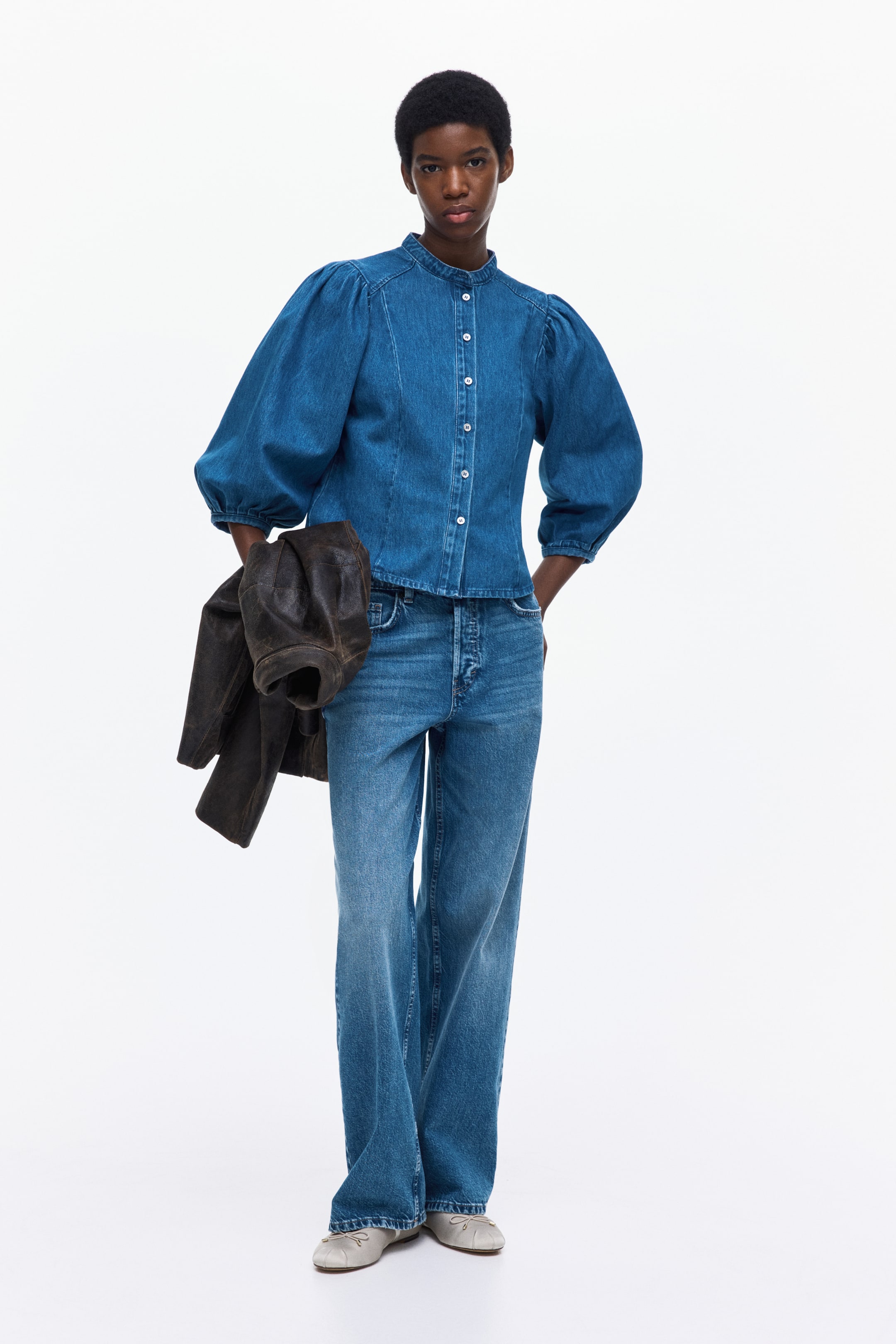 Grotere afbeelding bekijken: Een blauw denimhemd is frontaal afgebeeld, met een bandkraag en volumineuze pofmouwen die bij de schouders gerimpeld zijn en boven de pols eindigen. Het is voorzien van een volledige knoopsluiting aan de voorkant met lichtgekleurde knopen en verticale vormgevende naden op het lijfje.