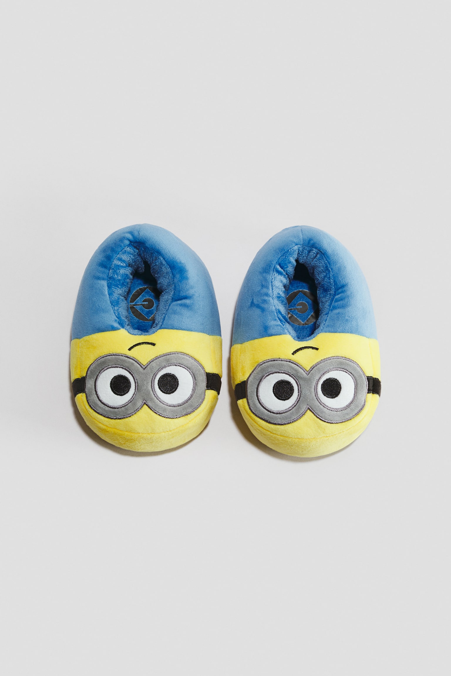 A pair of padded fabric slippers featuring a Minion character design is resting side by side, facing forwards. The slippers showcase a yellow front section with embroidered goggle and eye details, transitioning to a blue upper section with a plush, lined interior.