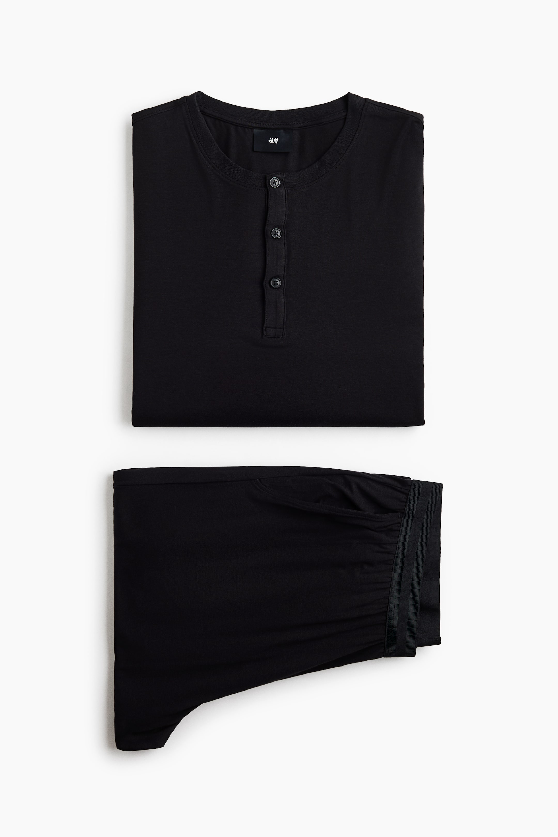 Größeres Bild ansehen: Ein schwarzes Pyjama-Set liegt flach gefaltet da und zeigt ein Langarmoberteil mit Rundhalsausschnitt und einer Drei-Knopf-Leiste, platziert über der passenden Hose mit elastischem Bund und einer sichtbar diagonalen Seitentasche.
