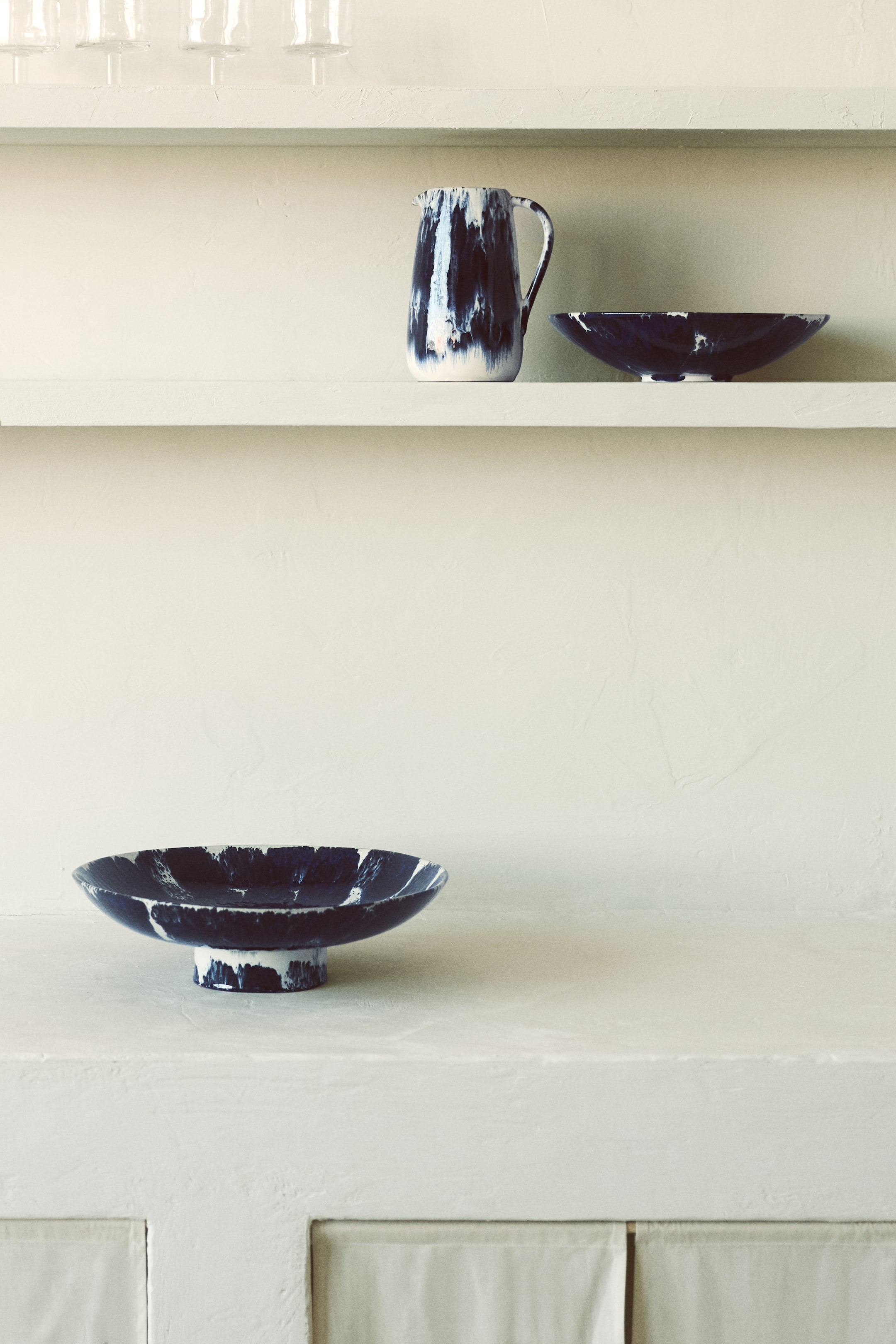 Ver imagem maior: Uma coleção de peças de servir em cerâmica é apresentada, com um design marmoreado em azul escuro e branco. Um jarro com pega e bico está posicionado na vertical, ligeiramente inclinado para a esquerda, ao lado de uma taça rasa com pé, repousando com a sua ampla abertura visível. Abaixo, uma taça maior e mais profunda é exibida sobre uma base cilíndrica em pedestal, também ligeiramente inclinada para a esquerda.
