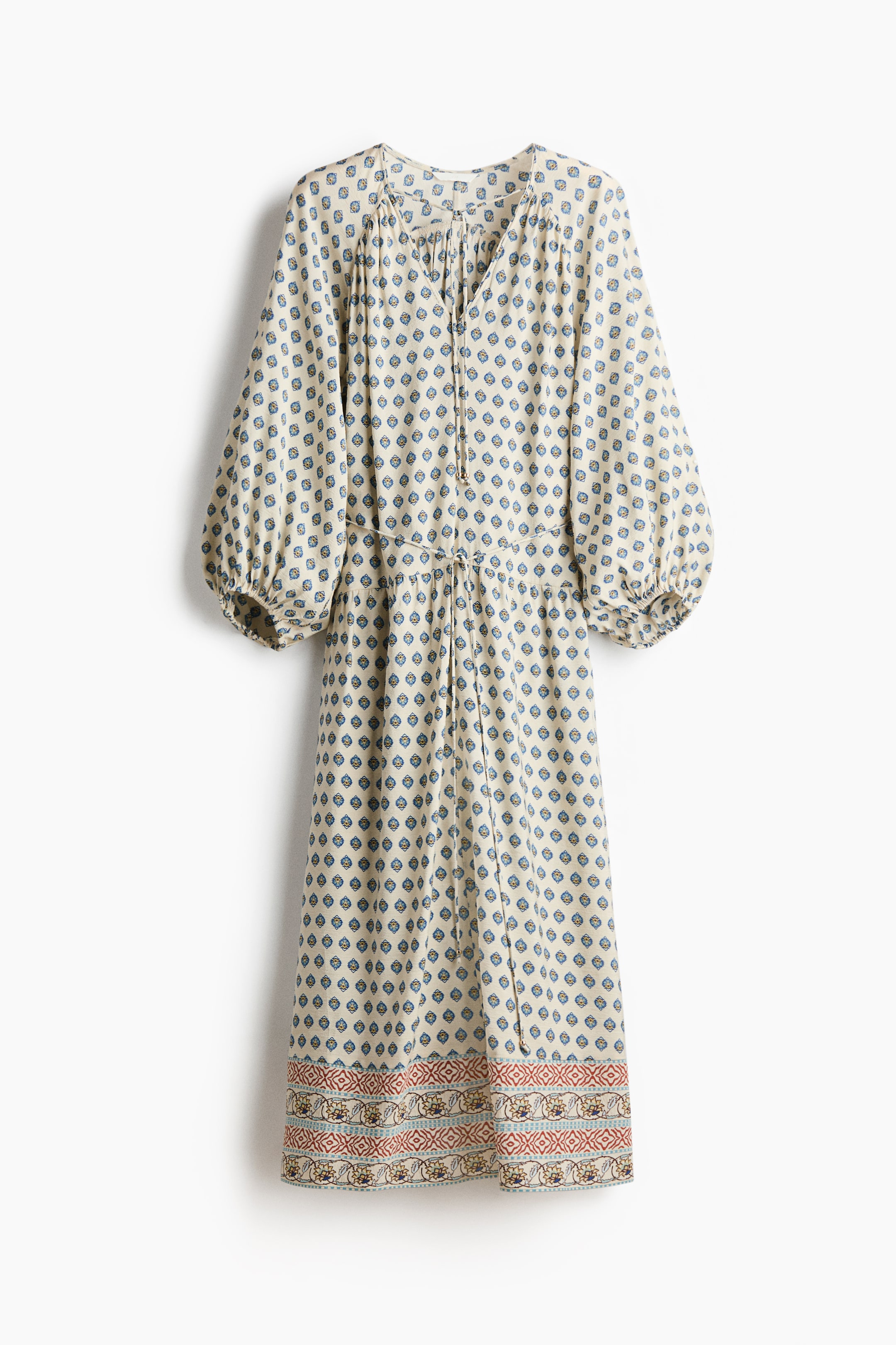 View larger image: A cream-coloured dress featuring a repeating blue and brown geometric pattern, a V-neckline with ties, and voluminous long sleeves with elasticated cuffs. It displays a drawstring waist and a wide decorative border with red, blue, and yellow patterns along the hem.