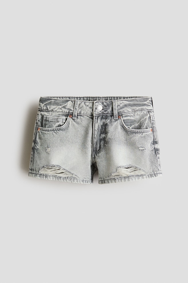 Estos shorts de 5 bolsillos presentan un ajuste relajado en denim de algodón gris lavado al ácido, vistos desde el frente. Cuentan con una pretina ajustable y elástica con cierre de cremallera y botón, detalles desgastados y rasgaduras en el frente, y dobladillos sin rematar. - 1
