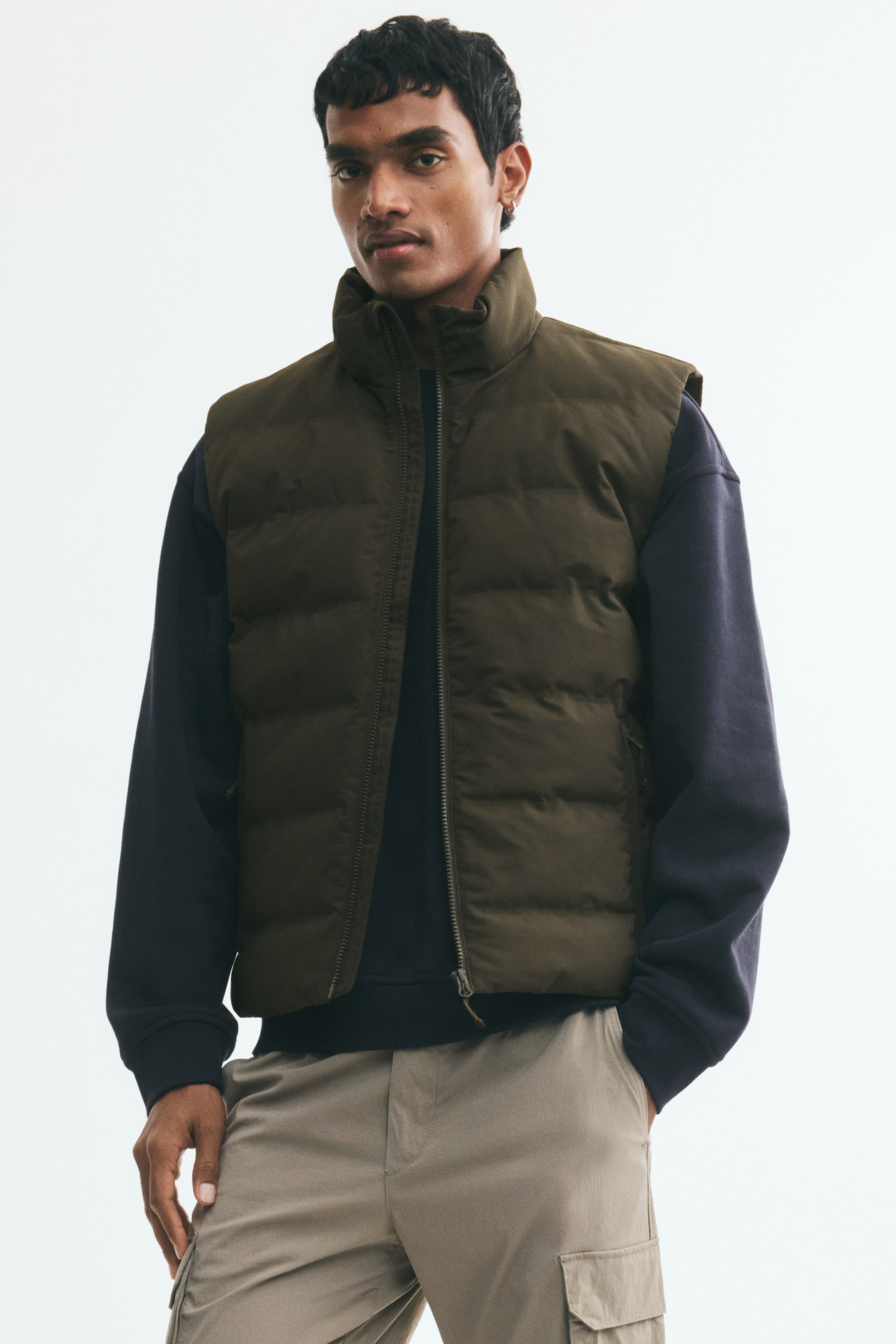 View larger image: The model is wearing an olive-green quilted vest with a stand collar and a visible front zipper. It features subtle zipped side pockets and is layered over a dark long-sleeved top.