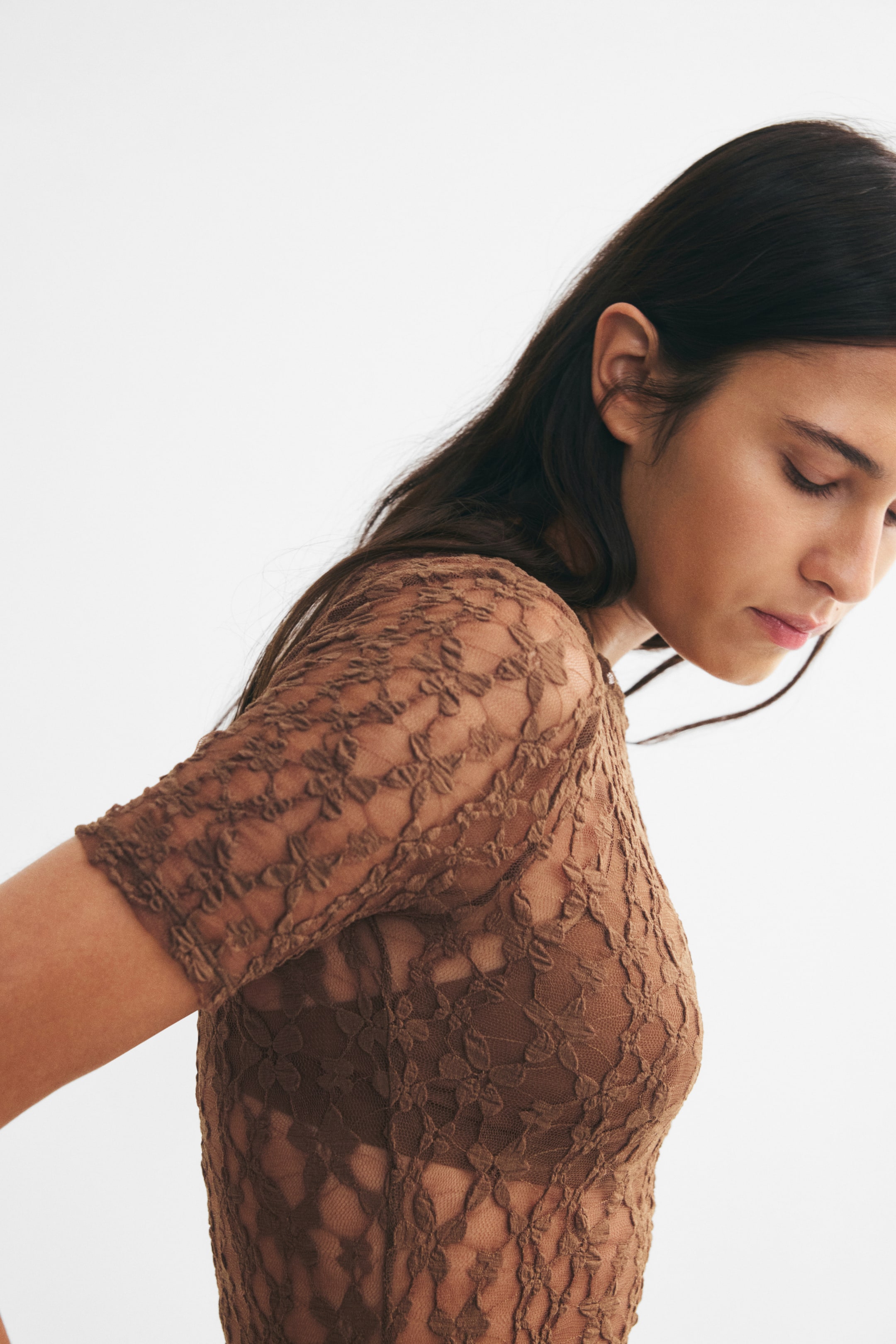 View larger image: The model is wearing a brown lace bodysuit with short sleeves and a floral pattern. The sheer lace fabric is layered over a matching brown bra, creating a fitted silhouette with a round neckline.