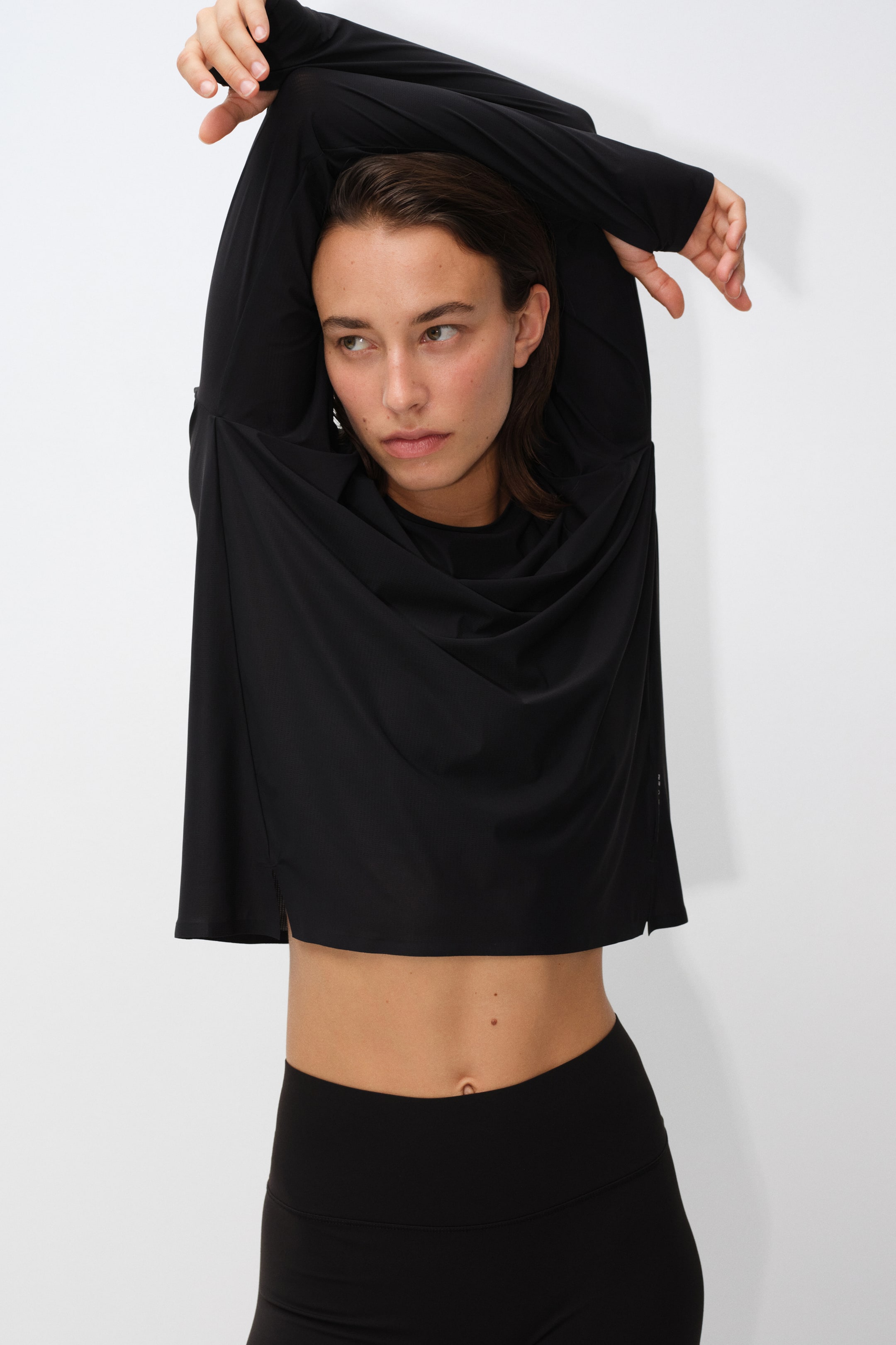 Visualizza immagine più grande: La modella indossa un top corto nero, ampio e a maniche lunghe, con le braccia sollevate sopra la testa, facendo drappeggiare il tessuto leggero sul petto. Il top è abbinato a leggings neri a vita alta coordinati.