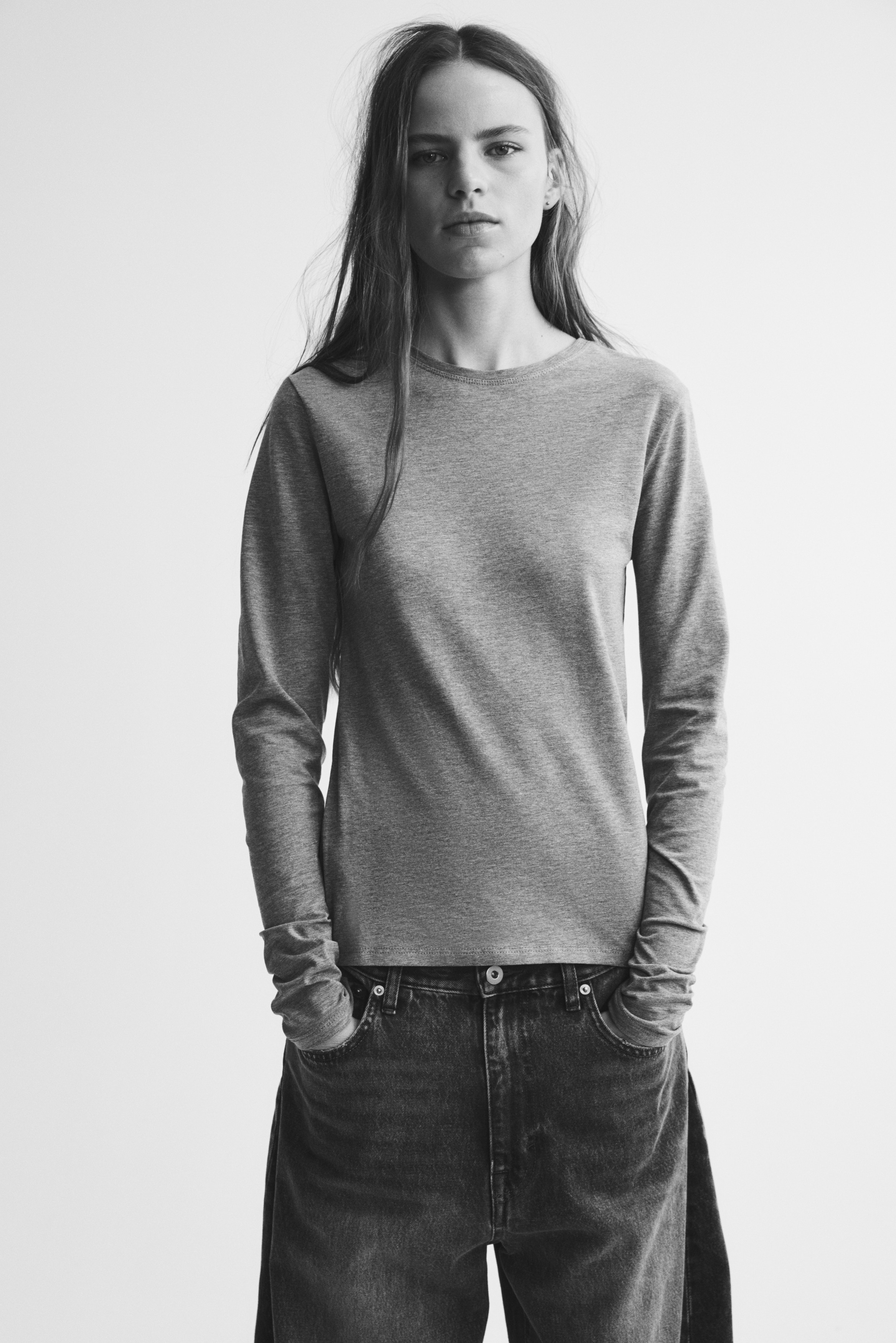 Ein graues Langarm-Top hÃ¤ngt mit der Vorderseite nach vorn und zeigt einen Rundhalsausschnitt, eine figurbetonte Silhouette und einen geraden Saum. Das weiche Baumwoll-Jersey-Material weist ein hellgraues Meliermuster mit schlichten gesteppten AbschlÃ¼ssen auf.