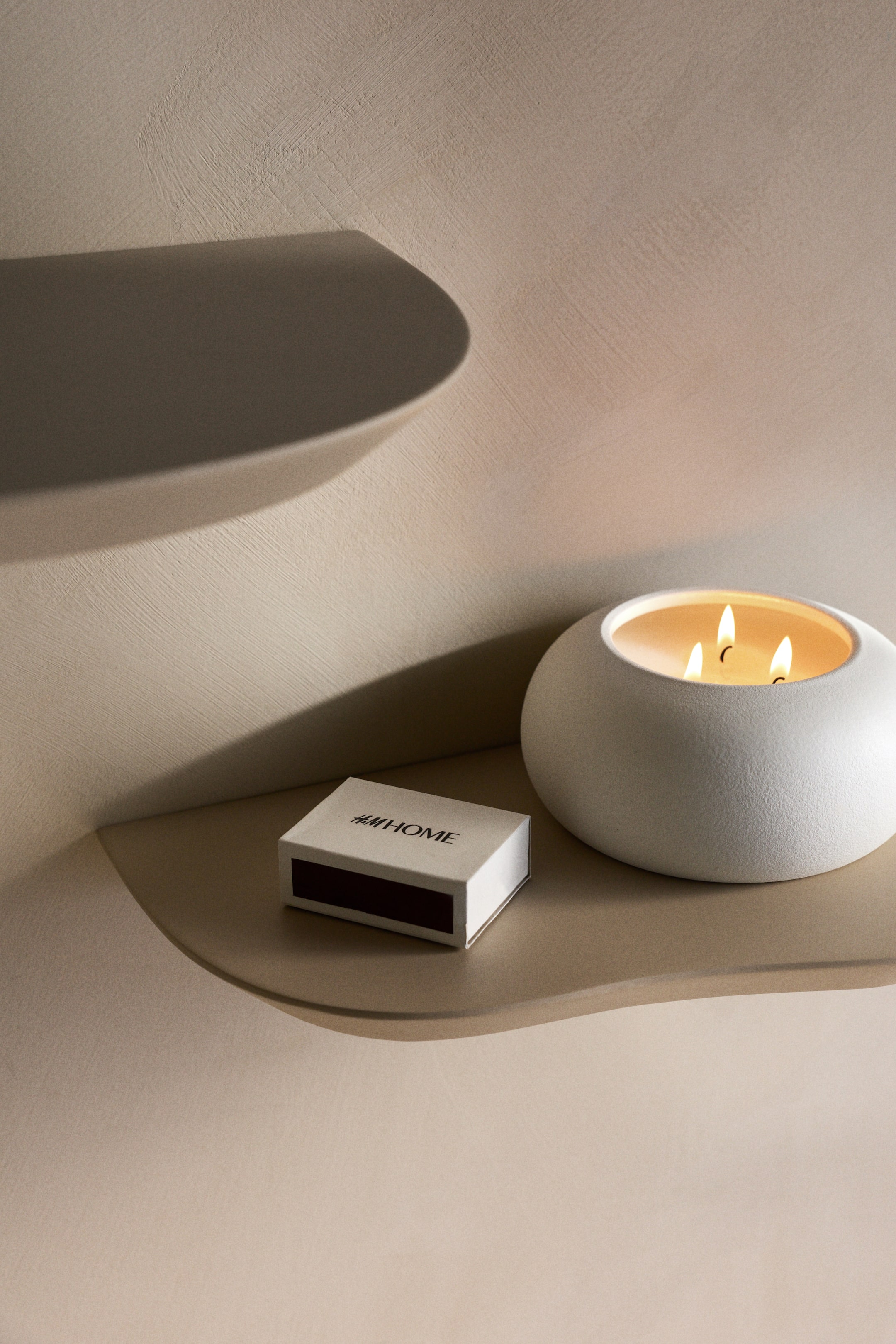 Ver imagem maior: Uma vela branca, texturizada, em forma de taça com três pavios acesos está ao lado de uma caixa de fósforos branca com a inscrição "H&M HOME."