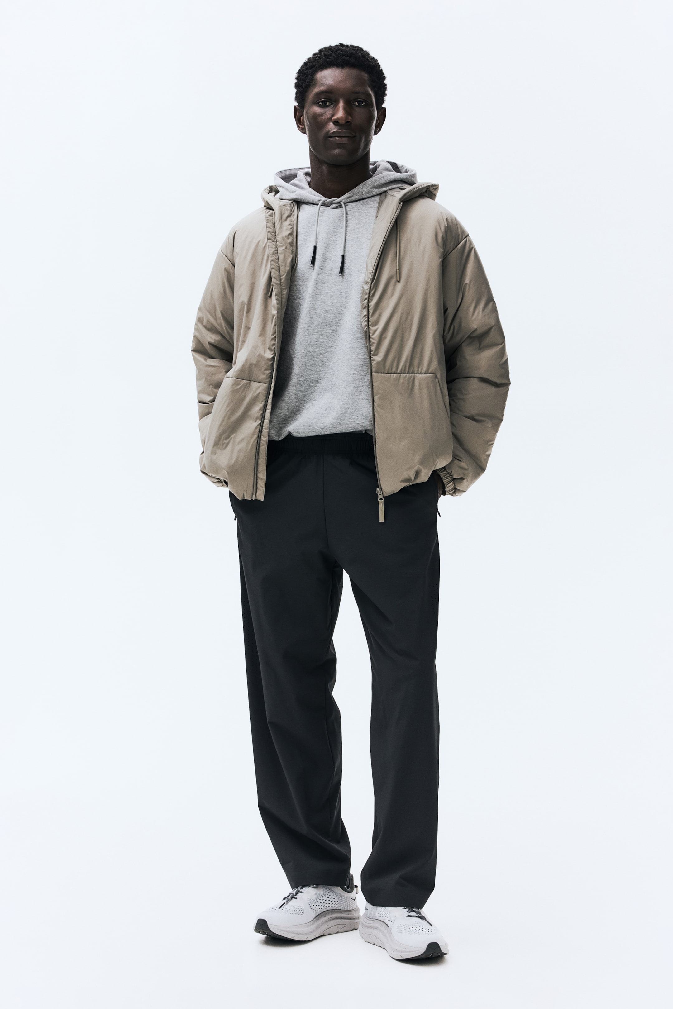 Voir une image plus grande: La tenue présente une veste matelassée beige clair à fermeture à glissière sur toute la longueur et munie d'une capuche, portée par-dessus un sweat à capuche gris clair avec cordons apparents. Un pantalon noir à coupe décontractée, doté d'une taille élastique et d'une poche latérale zippée, est porté avec des chaussures de sport blanches et gris clair.