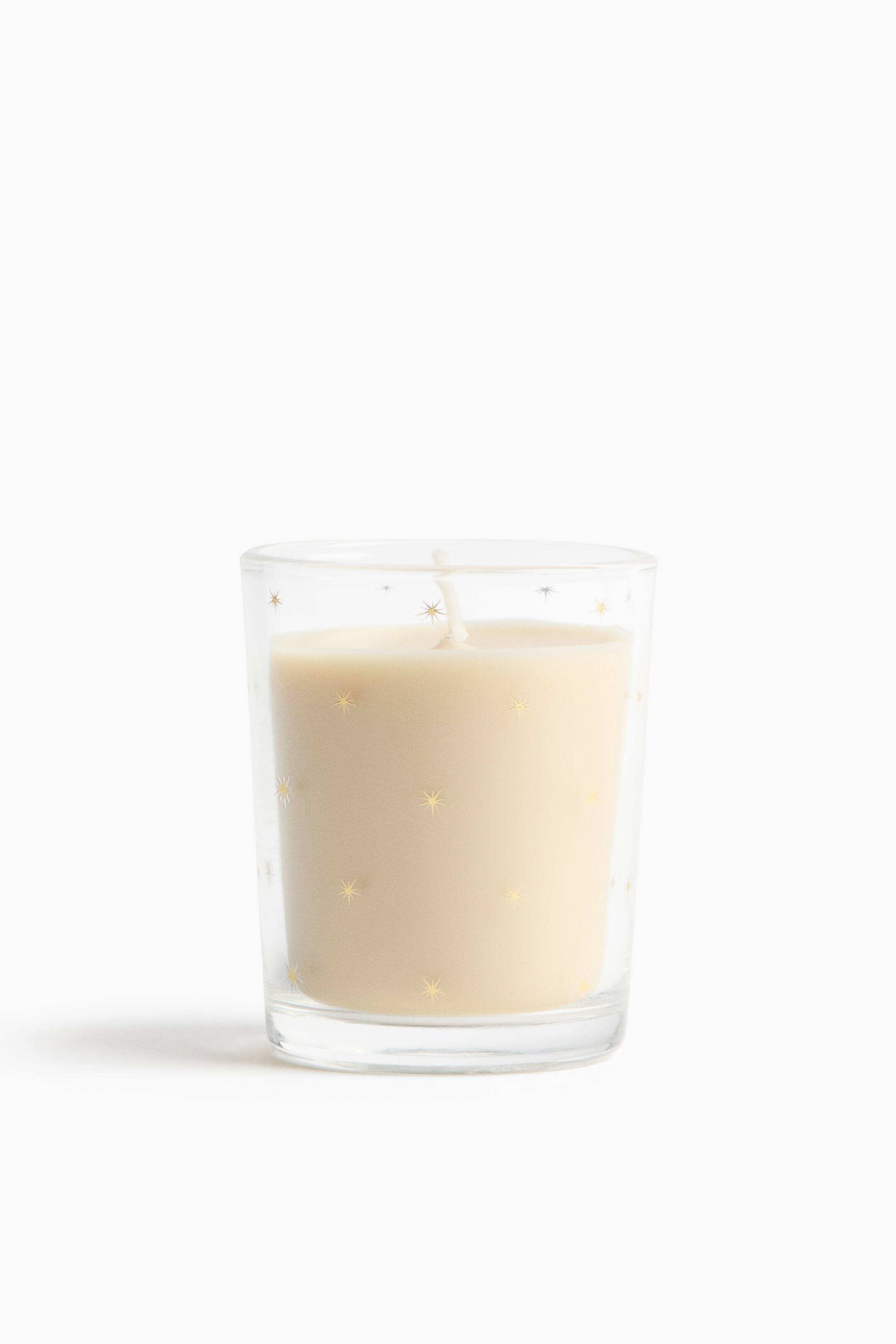 View larger image: A cream-coloured wax candle is resting inside a clear, front-facing glass tumbler featuring a pattern of scattered small gold and silver starbursts. A single white wick emerges from the centre of the smooth wax surface.