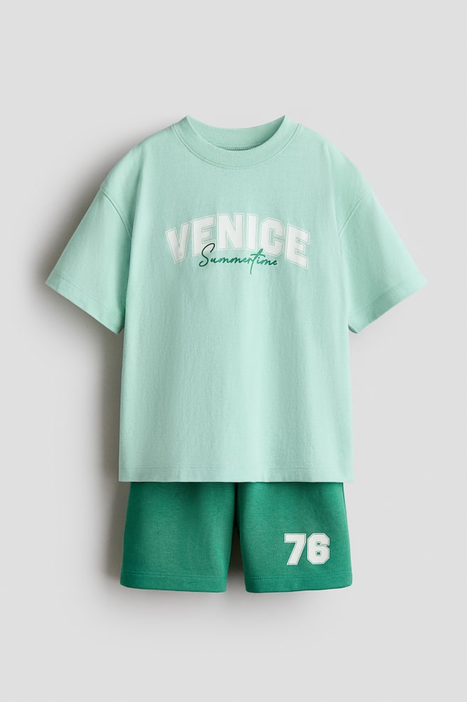 A two-piece set is presenting a light green T-shirt and dark green shorts, viewed from the front. The soft cotton jersey T-shirt is featuring "Venice" in an arched block font and "Summertime" in a script font on the front, with a ribbed neckline and dropped shoulders. The lightweight sweatshirt fabric shorts are displaying the number "76" on the left leg and are designed with an elasticated, drawstring waist. - 1