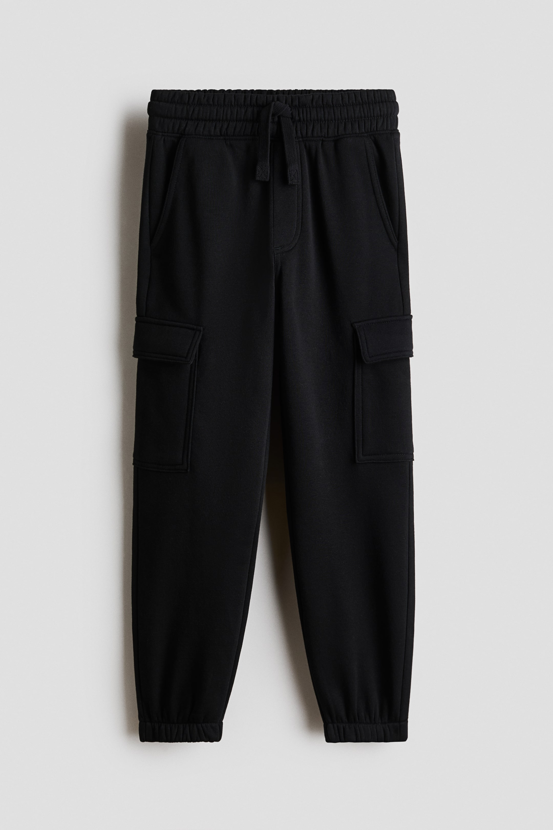 View larger image: Black cargo joggers are shown from a front view, featuring an elastic waistband with a visible drawstring tie. Two side pockets are located at the hips, and two rectangular flap pockets are positioned on the outer thighs, while elastic cuffs gather at the ankles.