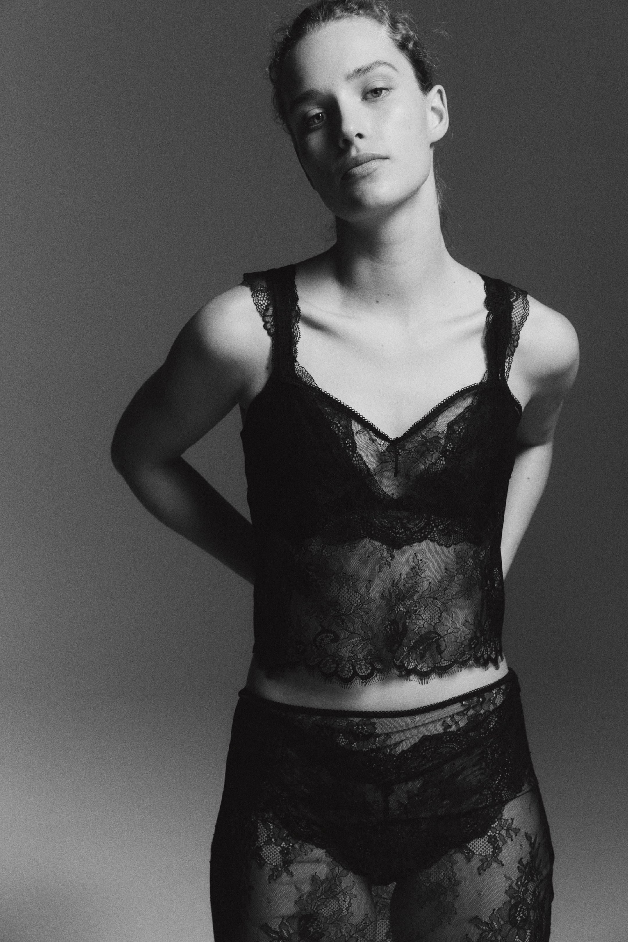 Se større billede: Et sort blonde lingerisæt med en camisole-stil top, der har bølgede blondestropper og en V-udskæring, der fremhæver gennemsigtig blomsterblonde med en bralette-stil uigennemsigtig panel ved busten. Matchende højtaljede trusser præsenterer gennemsigtig blomsterblonde med en uigennemsigtig foring, hele sættet sidder tæt til kroppen.