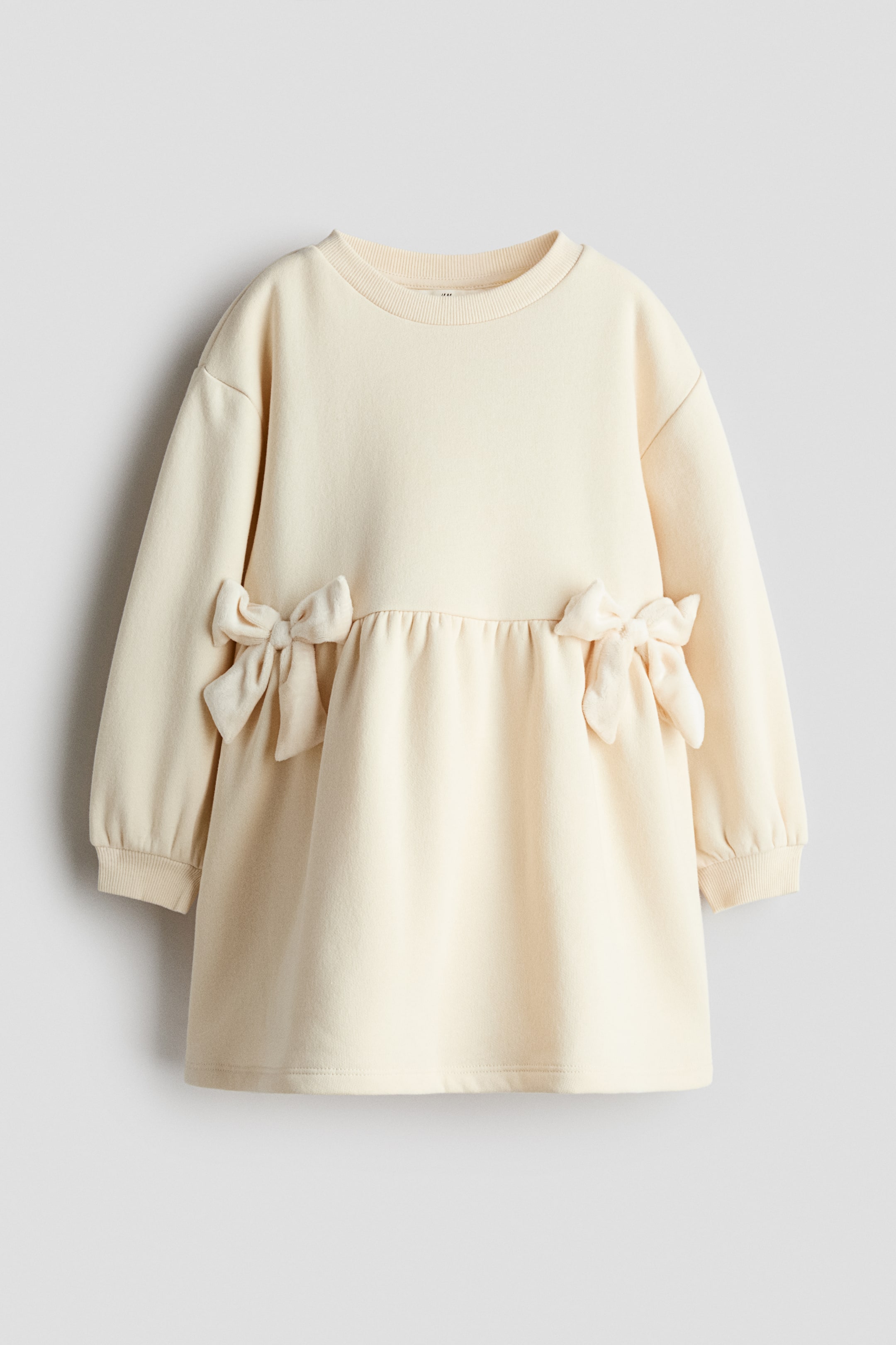 View larger image: A cream-colored dress made from brushed fleece fabric is shown from the front, featuring a round rib-trimmed neckline, dropped shoulders, and long sleeves with ribbed cuffs. A gathered seam at the waist is accented by a decorative velour bow on each side, leading into a flared skirt.